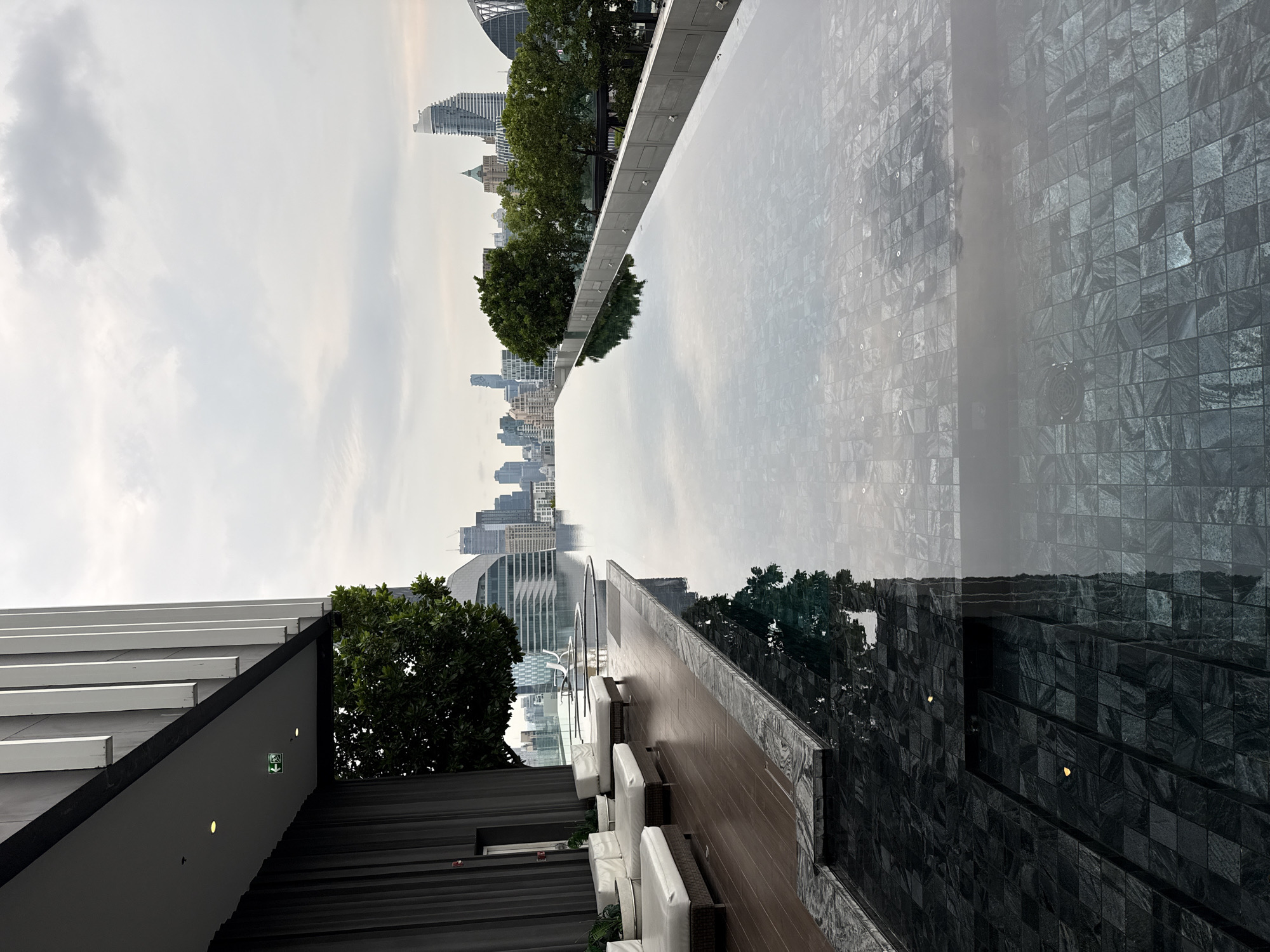 A rooftop pool scene in Bangkok, Thailand, presents a stark contrast between urban skyline reflections and architectural detail. The pool's surface mirrors the city's tall buildings and cloudy sky, bisecting the frame with a horizon line. Below, a wooden deck lined with white cushioned lounge seating and green foliage faces the cityscape. The pool's edge is constructed with dark, mosaic-tiled walls, leading to an undefined depth. A modern, angular building's overhang is visible in the lower left corner, marked with safety lighting.