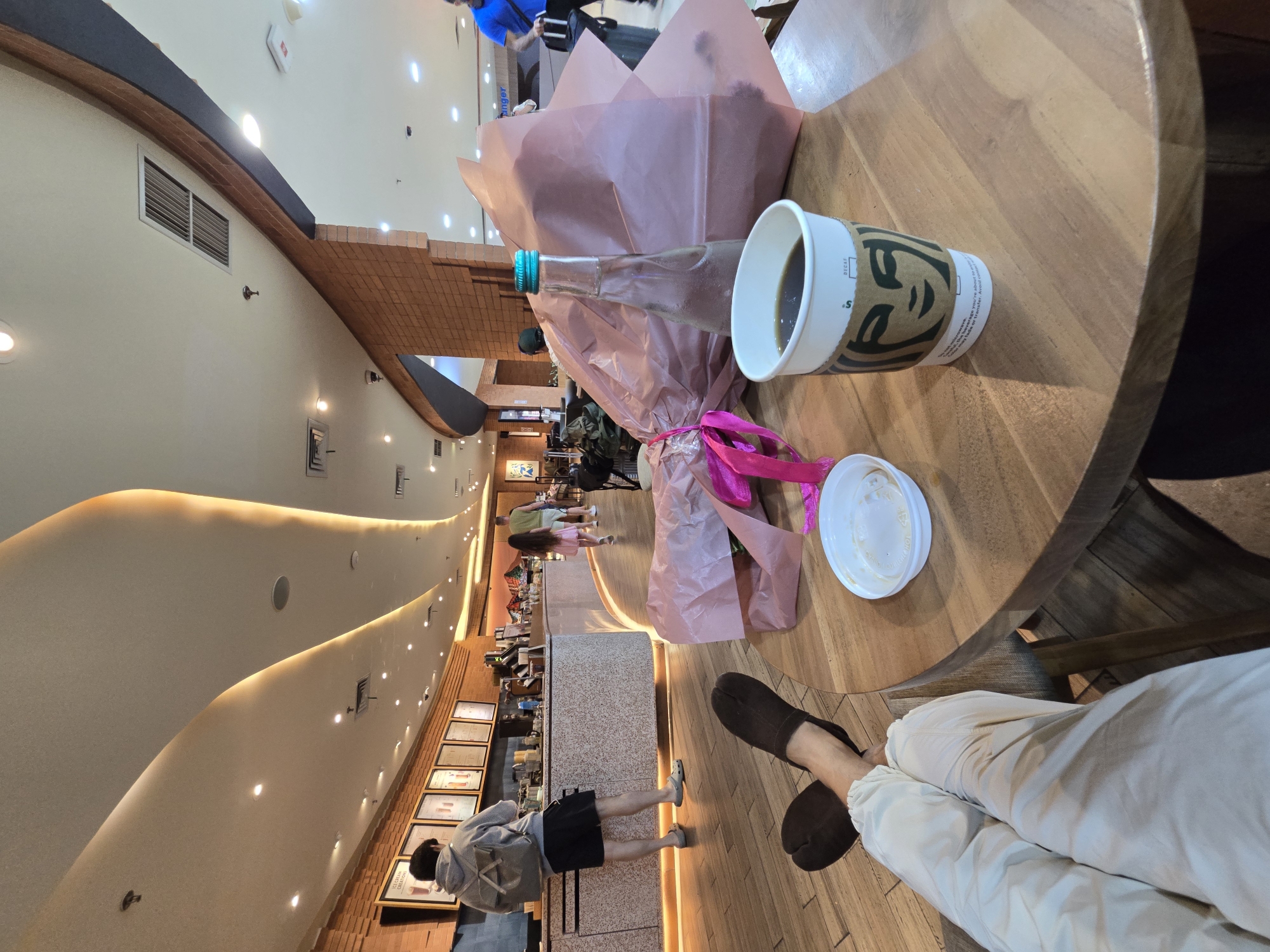 This media file captures an indoor scene at a cafe or dessert shop, likely in Kuta, Indonesia. The image is taken from a first-person perspective, showing a person's legs dressed in light-colored pants and dark, possibly traditional, slip-on shoes, resting beneath a round wooden table.
On the wooden table, there's a white Starbucks coffee cup with a brown sleeve, a glass water bottle with a blue cap, and a large item wrapped in pink paper, secured with a bright pink ribbon, possibly a gift or bakery item. An empty white cup lid is also visible. The coffee cup sleeve clearly shows text including "DECAF", "microwave.", and a partial warning: "beverage you're about to transfer. Avoid conte de or tre".
The background reveals a bustling cafe environment. A long service counter runs through the midground, with display cases and framed items. Several patrons are visible; one person in a grey hoodie and black shorts stands at the counter, looking at framed pictures, while others are further back, including a person in a pink dress. The architectural design features prominently, with curved, cream-colored walls illuminated by warm, recessed lighting and spotlights. Sections of the wall are adorned with brick-like textures and dark accents. Overhead, air vents are visible. Signs near the counter display the text "ICE CREAM CREATIONS" and a partial word "anger". The floor appears to be made of light-colored wood planks. The lighting suggests an indoor setting, likely during the day or early evening.