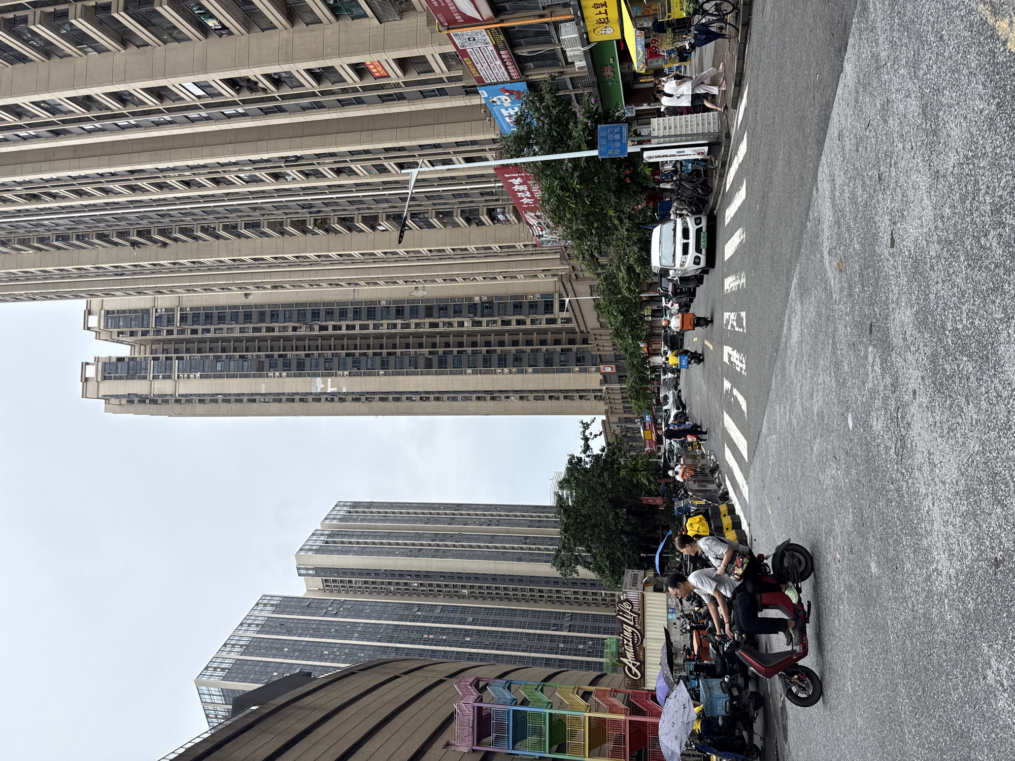 In Haizhu District, China, a city street is depicted with tall, modern residential buildings dominating the skyline. The buildings are characterized by their beige facades and numerous balconies. In the foreground, a man and a woman ride on a red electric scooter, traveling towards the left side of the image. The man is driving, and the woman is a passenger behind him. To the left of the scooter, parked delivery vehicles and umbrellas are visible. Across the street, pedestrians are walking on the sidewalk. The street itself is paved with asphalt and features a pedestrian crossing. The sky is overcast, suggesting a cloudy day. The scene captures a slice of urban life with a focus on transportation and the built environment.