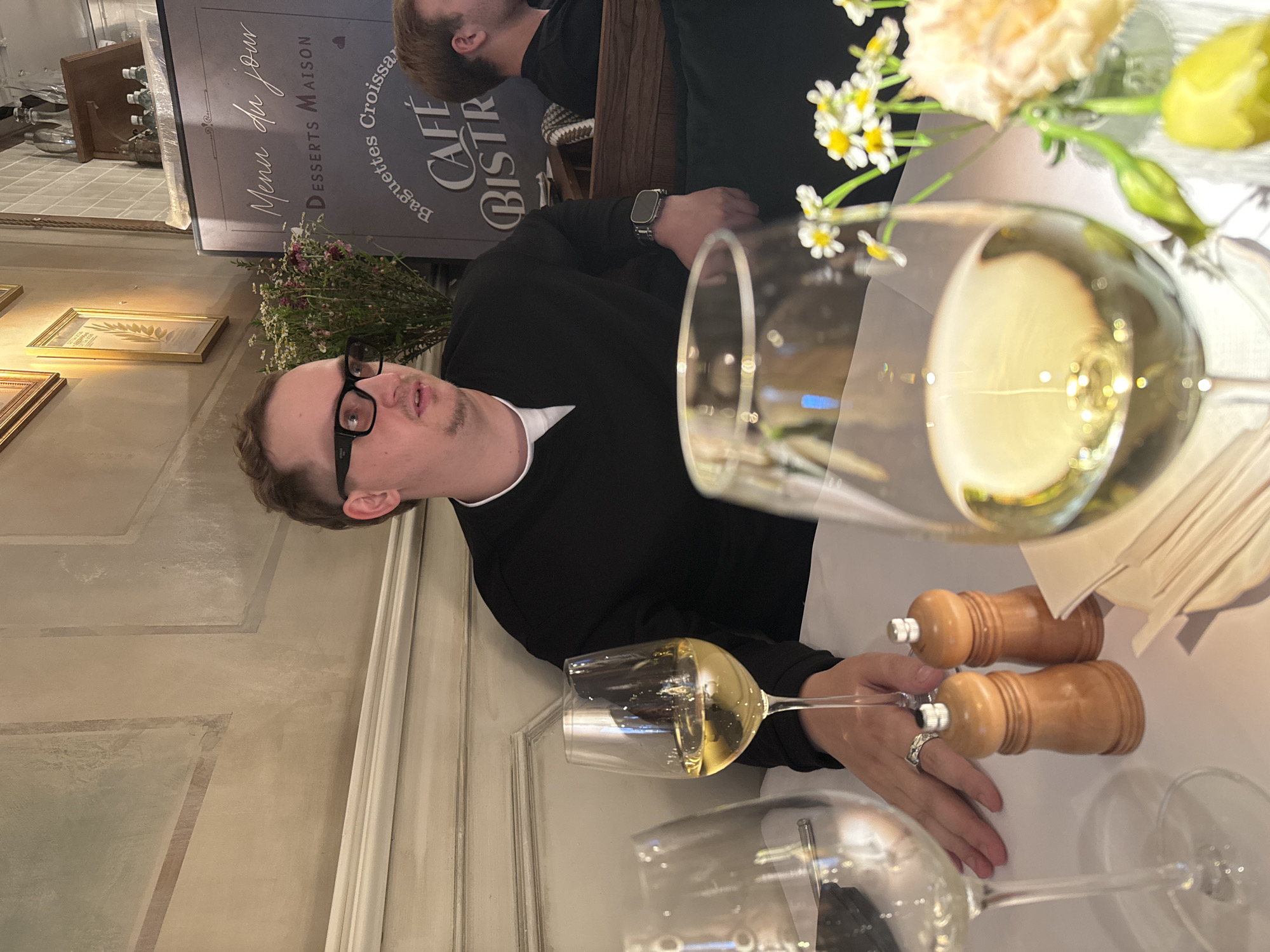 The image depicts a man sitting at a table in a cafe, likely in Moscow, Russia, judging by the signage which includes "Cafe Bistro" and "Menu du jour". He is wearing glasses and a black sweater, and has a smartwatch on his left wrist. In front of him, there are two glasses of white wine and a salt and pepper shaker. A bouquet of small white and yellow flowers is also visible on the table. The background shows a wall with ornate detailing and framed pictures. Another person, partially visible, is seated to the right of the main subject. The overall ambiance suggests a casual dining setting.