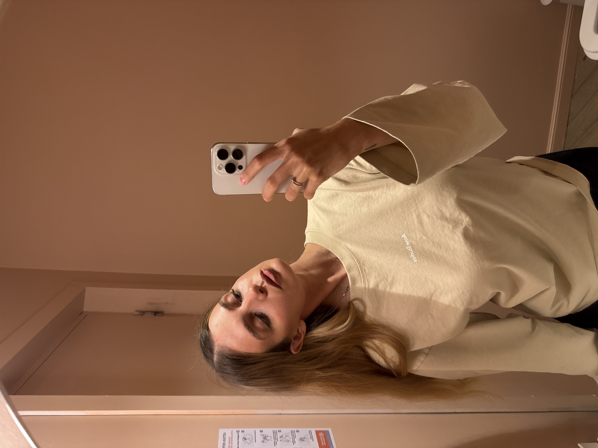 A selfie image shows a young woman holding a smartphone in a bathroom with peach colored walls. The woman has long, light brown hair and is wearing a light tan sweatshirt with a loose fit. She is wearing a ring on her left hand. A small poster is visible on the lower left of the wall. The location appears to be a bathroom or restroom. Information about the location in Tbilisi, Georgia is not available in the image.