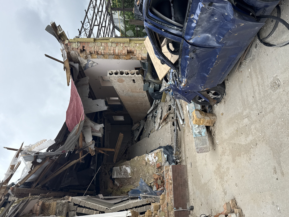 A damaged blue car rests against a partially collapsed brick building. The building's facade is severely compromised, revealing exposed interiors, including a white ceiling and concrete slabs. Debris litters the ground, alongside wooden planks and roofing materials. The sky is overcast. The scene conveys a sense of destruction, possibly from an impact or explosion.