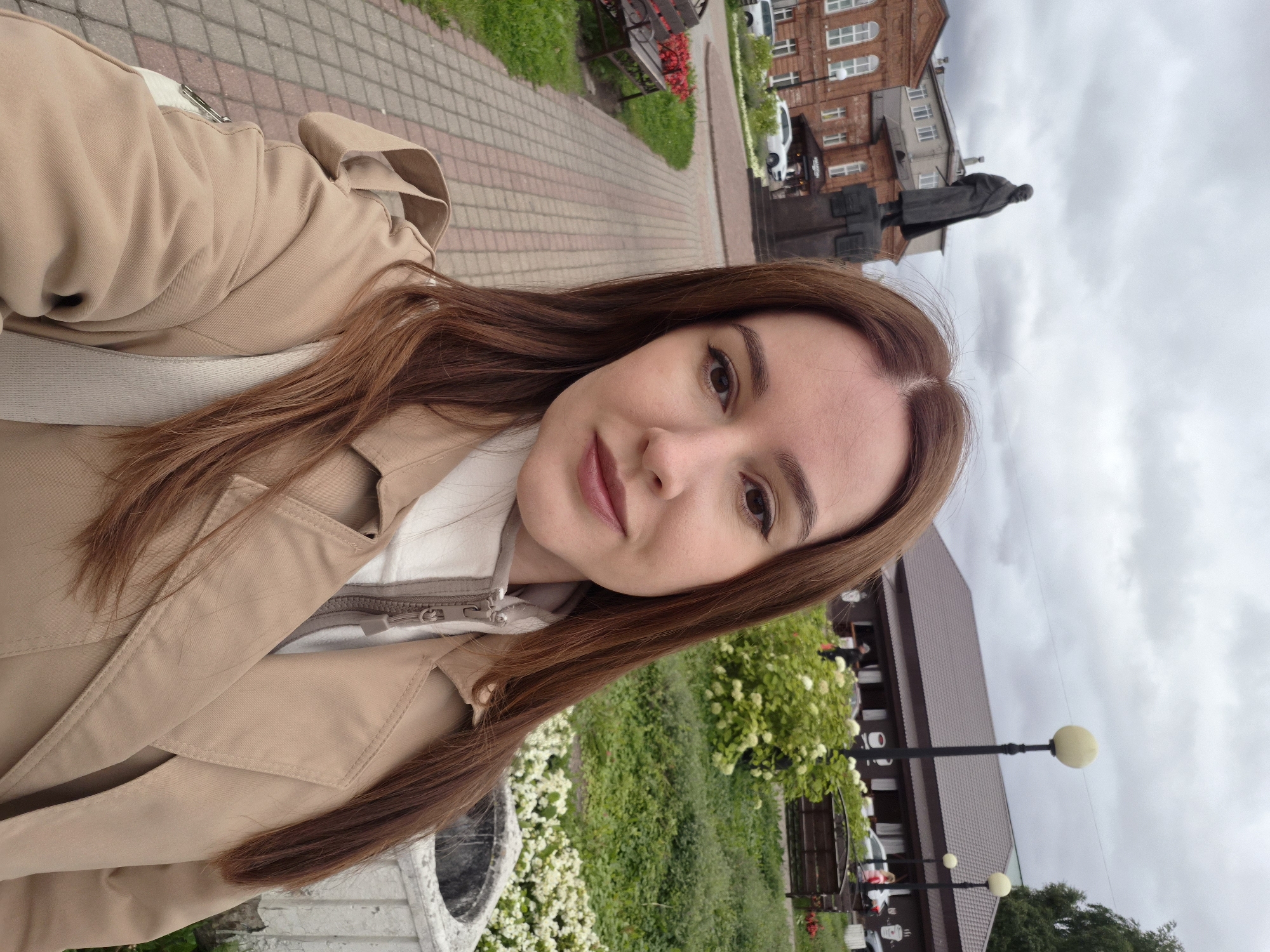 The image is a selfie of a young woman with long, brown hair and fair skin, wearing a beige trench coat over a white zip-up jacket. She is looking directly at the camera with a neutral expression.
The background suggests an outdoor urban setting. To the left, a paved walkway with reddish-brown bricks leads away from the viewer. Beside the walkway, there is a metal bench with red flowers in a planter. Further back, a large brick building with white-trimmed windows is visible, and in front of it stands a dark statue of a man in a long coat.
To the right of the walkway, there is a green grassy area with bushes and white flowers. Beyond this, a brown, low-rise building with a sloped roof is situated. Several lampposts with round, yellow lights are spaced along the front of this building. Cars are parked in front of the building, and a few people can be seen in the distance. The sky is overcast with gray clouds.
The image was taken on August 27, 2023, at 12:32 PM, in Cherepovets, Russia. The location is specifically identified as Lenin Street, near the intersection with Oktyabrsky Boulevard. The statue in the background is a monument to Vladimir Lenin. The building to the right is likely a cafe or restaurant, suggested by the cups depicted on its facade.