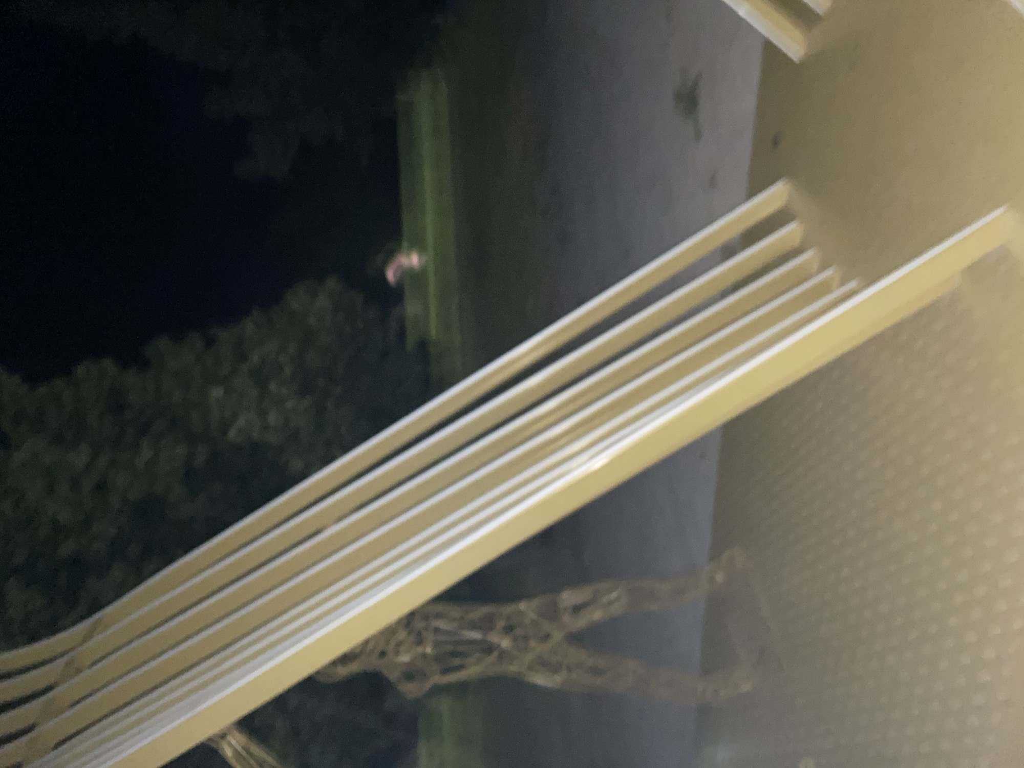 At night, an abstract, human-shaped sculpture made of wire mesh stands on a paved surface in what appears to be a public park or playground. The sculpture is located to the left of the frame, partially obscured by a structure with yellow and white parallel bars, possibly a slide or a railing. The ground is a mottled gray and beige. In the background, out of focus, there are dark trees and a patch of illuminated green grass. Further in the background and to the right, a small, blurry, light pink or orange object is visible on the grass. The overall lighting suggests artificial illumination from an unknown source, casting long shadows and creating a dark, atmospheric scene. The location is Yegorlykskaya, Russia.