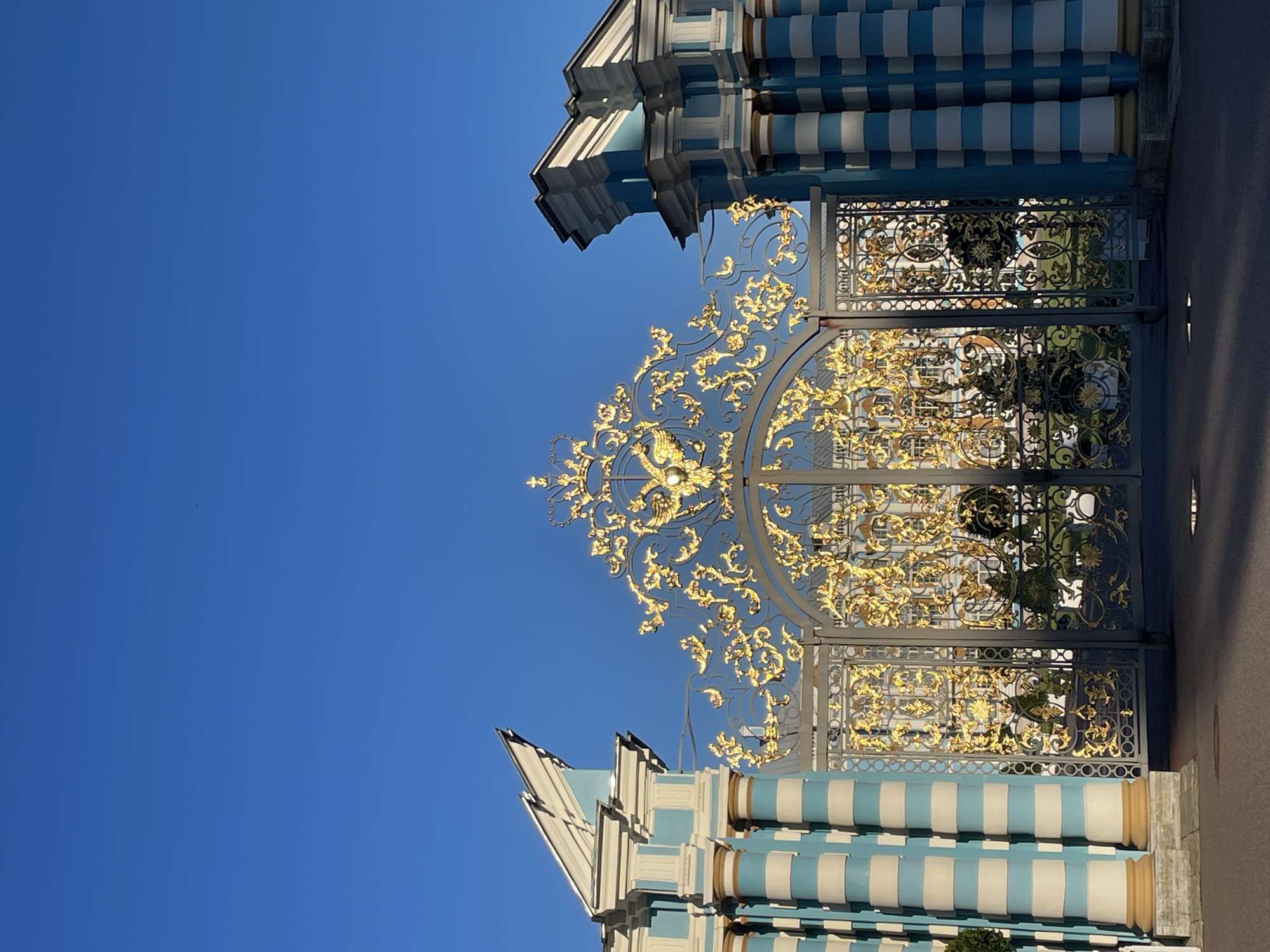 This image shows the Golden Gate of Catherine Palace in Pushkin, Russia. The gate is an ornate structure made of gilded metal with intricate scrollwork and a depiction of a double-headed eagle at the top. It is flanked by two pillared structures painted in alternating bands of light blue and white. The gate is set against a clear, bright blue sky. In the background, through the gate, a building with multiple windows and a mansard roof is visible, suggesting the presence of the palace itself. The ground in front of the gate is paved and casts shadows, indicating sunlight. There are no people or animals visible in the scene.