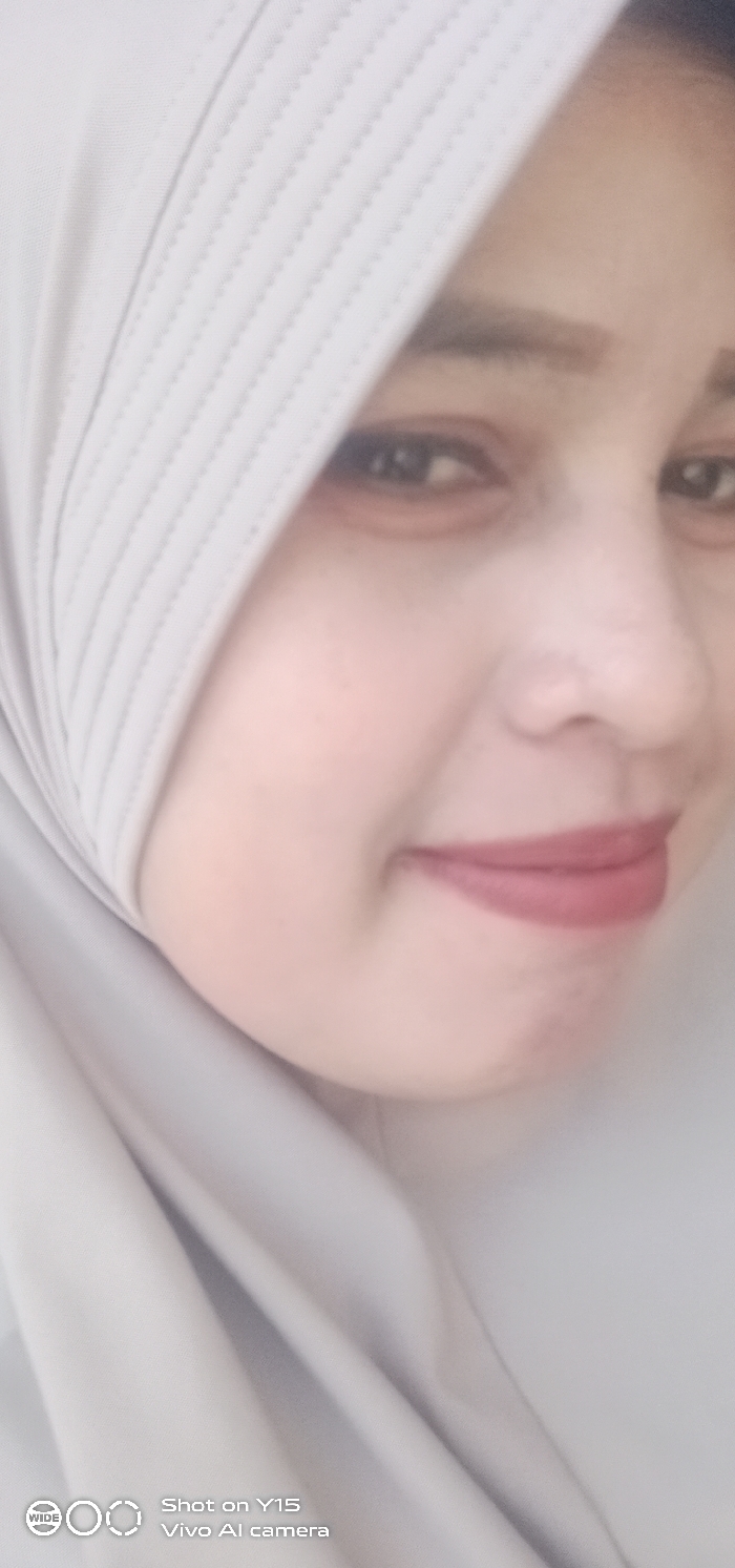 A close-up portrait shot features an adult female subject. Her face, oriented slightly to the right, occupies the majority of the frame, with her gaze directed forward. She displays a subtle smile, enhanced by reddish-brown lipstick on her lips. Her eyes are dark, complemented by dark, defined eyebrows. She is wearing a light grey or beige hijab, which covers her head and part of her shoulders; the portion over her forehead exhibits distinct vertical stitched patterns. The background is uniformly blurred and light grey/beige, providing no specific environmental context or identifiable structures. The location within the city of Gending, Indonesia, cannot be determined from the image's visual content. A digital watermark in the bottom-left corner indicates "Shot on Y15 Vivo AI camera". No other individuals, objects, or actions are depicted.