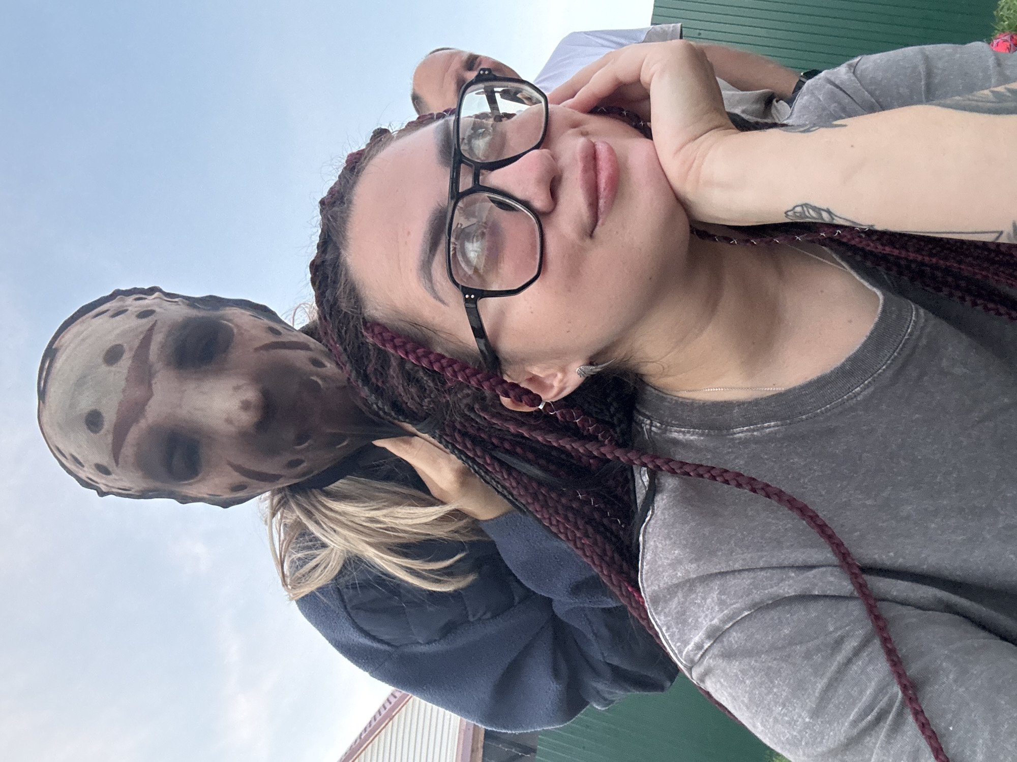 In a daylight outdoor scene, two individuals are prominently featured in the foreground. The individual closer to the camera, wearing a grey t-shirt and black-rimmed glasses, rests their chin on their hand. Their dark hair is styled in braids with red highlights. To the left and slightly behind, another person is visible, wearing a dark blue jacket and a mask resembling a hockey mask, with blond hair partially visible. In the background, another person wearing a white t-shirt can be seen. The setting includes a green fence and a building with a red roof. The sky is clear blue. The location is specified as Узюково, Russia.