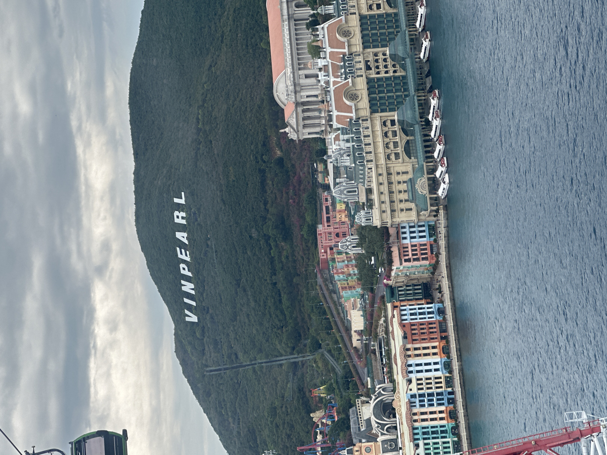 An aerial view shows Vinpearl, Nha Trang Ward, Vietnam, beside the sea. A green, forested mountain dominates the scene, with "VINPEARL" written in large, white letters on its slope. A series of buildings, designed in a European style, runs along the coast, transitioning from neutral tones to colorful pastels. Along the coast, multiple boats are parked. The sea is a shade of blue. The sky is overcast. A cable car structure is partially visible in the foreground. Amusement park rides can be seen at the base of the mountain, along with roads crossing through the mountain.