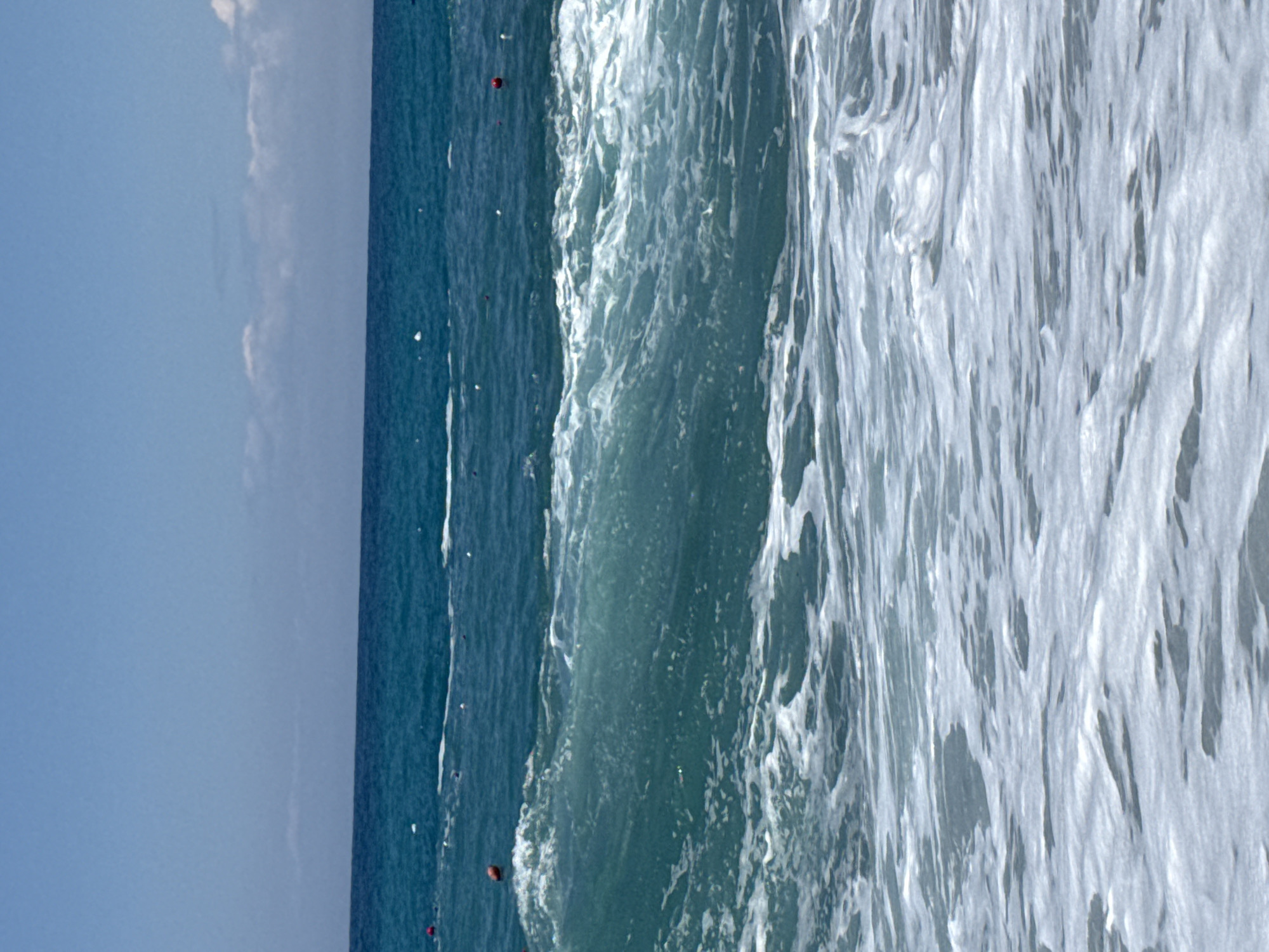 The image shows a seascape with a blue sky occupying the left side of the frame. The horizon line is visible, dividing the sky from the ocean. The ocean is predominantly blue with white foamy waves crashing in the foreground, moving from left to right. There are several small red buoys or markers visible in the water. The exact location is unknown.