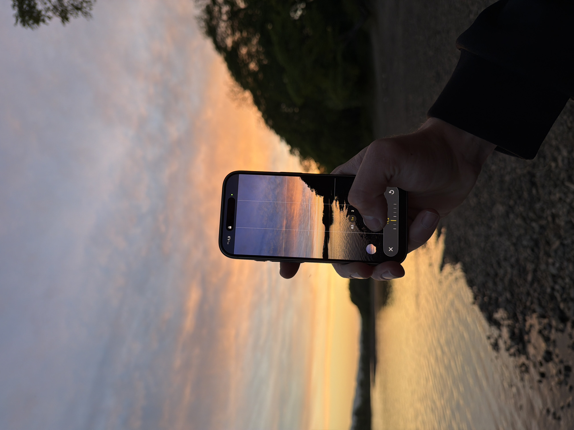 A person holds a smartphone up horizontally to take a picture of a sunset reflecting on water. The phone's screen displays a similar scene: a body of water stretching to the horizon where the sky transitions from shades of blue to orange and yellow. A dark landmass or forest is visible in the upper right quadrant of the phone screen and of the image. The person's hand and part of their arm, clothed in a dark garment, occupy the right side of the frame. The environment appears to be a shoreline, with the sky dominating the left side of the frame and the water on the right. It's unlikely to determine that this image was taken in Birobidzhan, Russia without location data.