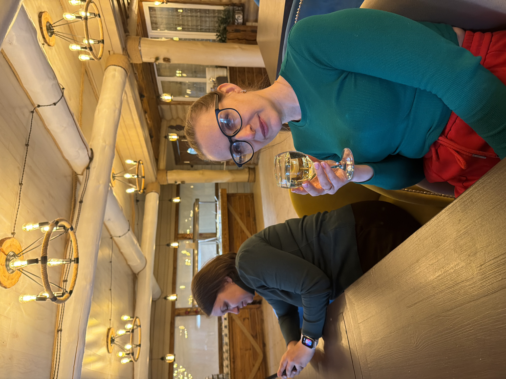 Two women are at a table in a building in Zaplav'e, Russia. The woman closer to the camera is holding a glass of white wine. She is wearing a green long-sleeved shirt, red pants, and glasses. The other woman is looking at a phone. She is wearing a green long-sleeved shirt and a watch. The background features lights, windows, and wooden support beams.