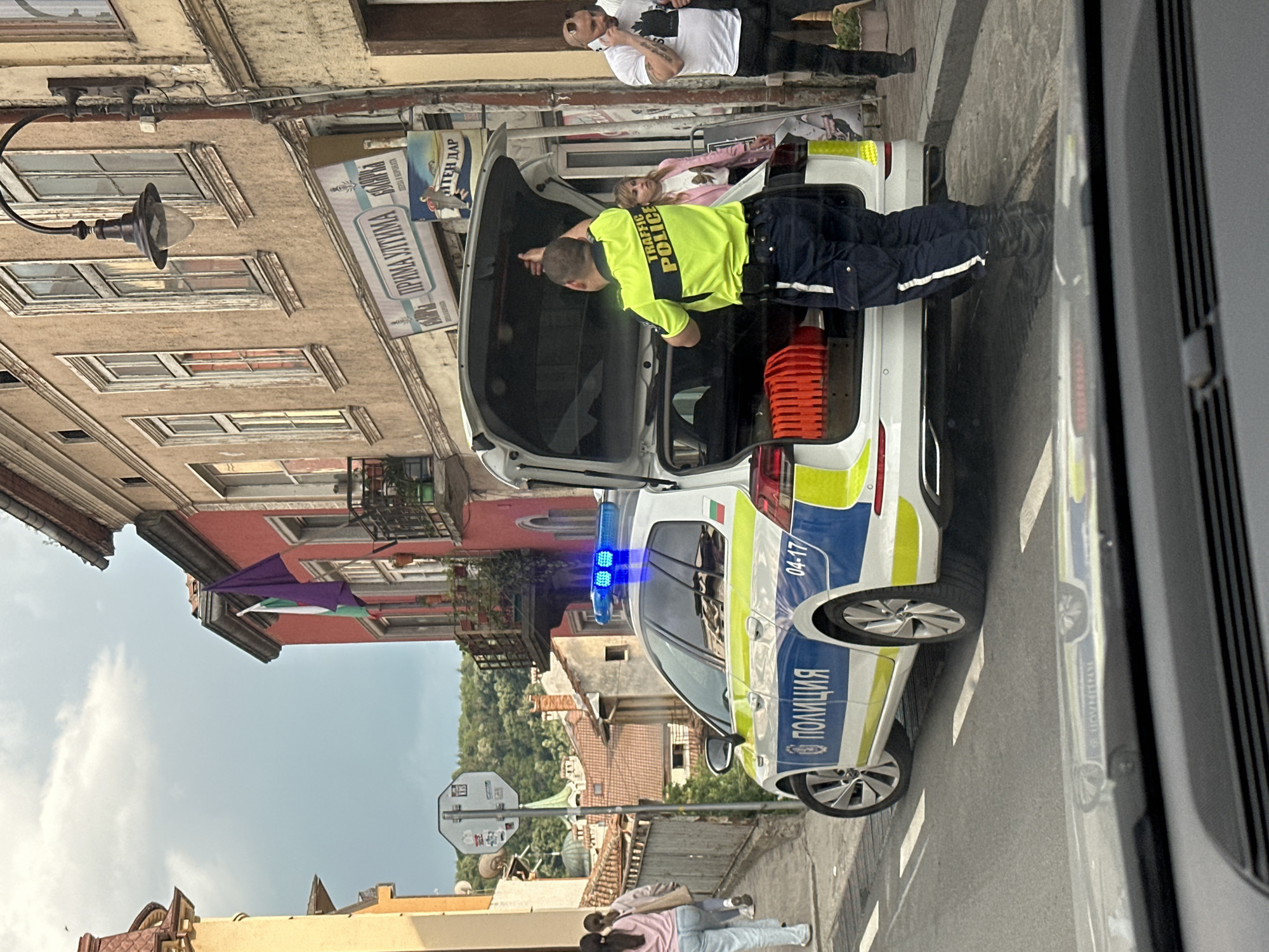 A street scene in Veliko Tarnovo, Bulgaria shows a police car with its hatchback open. A police officer in a bright yellow "Traffic Police" vest is looking into the car's trunk, where a young girl is lying down. A man in a white shirt is standing behind the car, talking on a cell phone. In the background, buildings characteristic of Veliko Tarnovo are visible, along with the Bulgarian flag on a building.