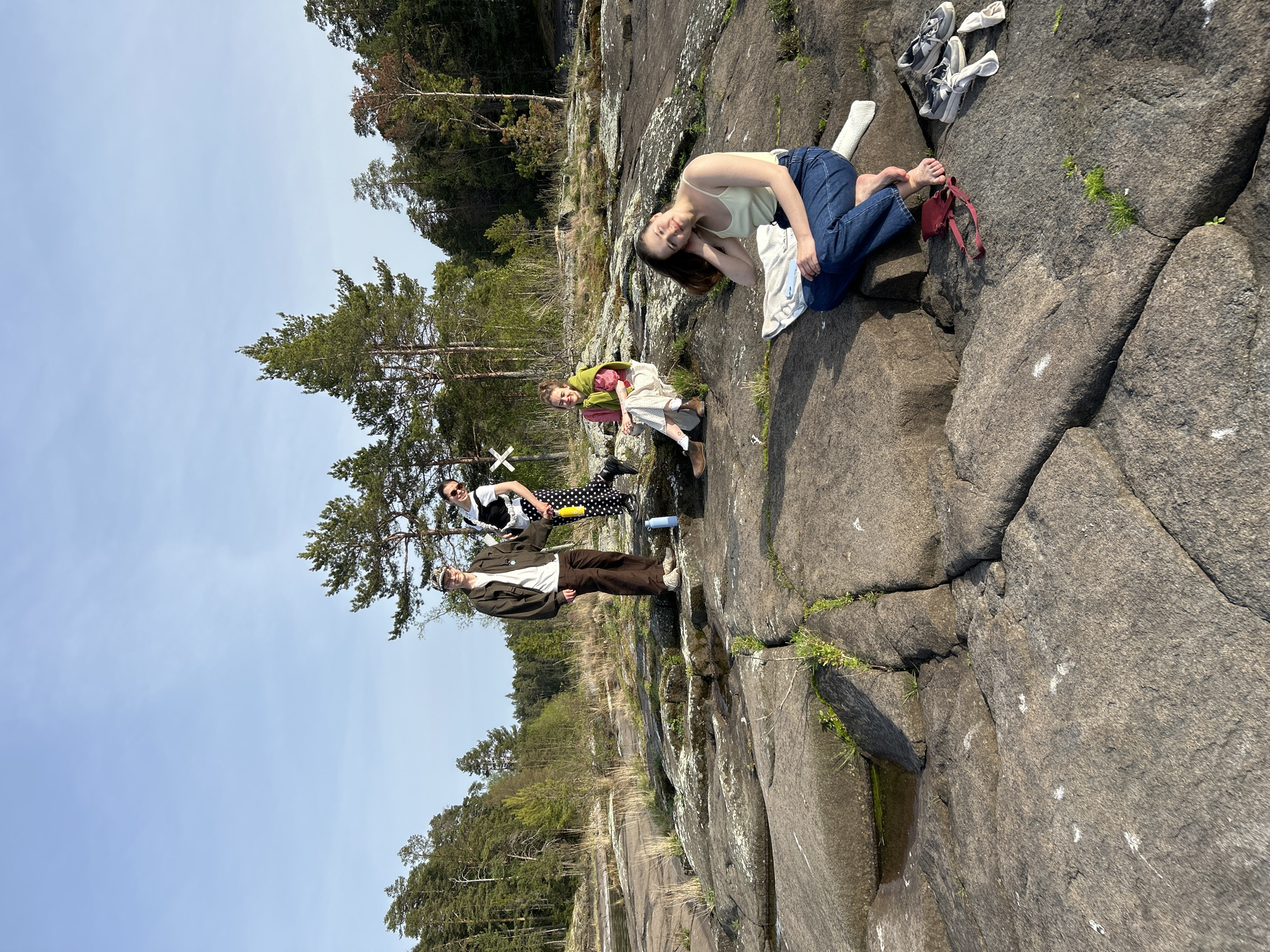 Four individuals are positioned on a rocky terrain near a body of water, possibly in Valaam, Russia. The terrain consists of large, gray rocks, some with patches of green moss or vegetation. The background features trees and a clear blue sky. A young woman kneels near the edge of the rocky area, wearing a light-colored tank top and jeans. She rests her head on her hand. Beside her are a pair of shoes and a red bag. A second woman sits behind her, wearing a patterned dress and a green shawl. Behind them stands a person with sunglasses and a black and white top. Further back stands a person wearing a brown jacket and pants.