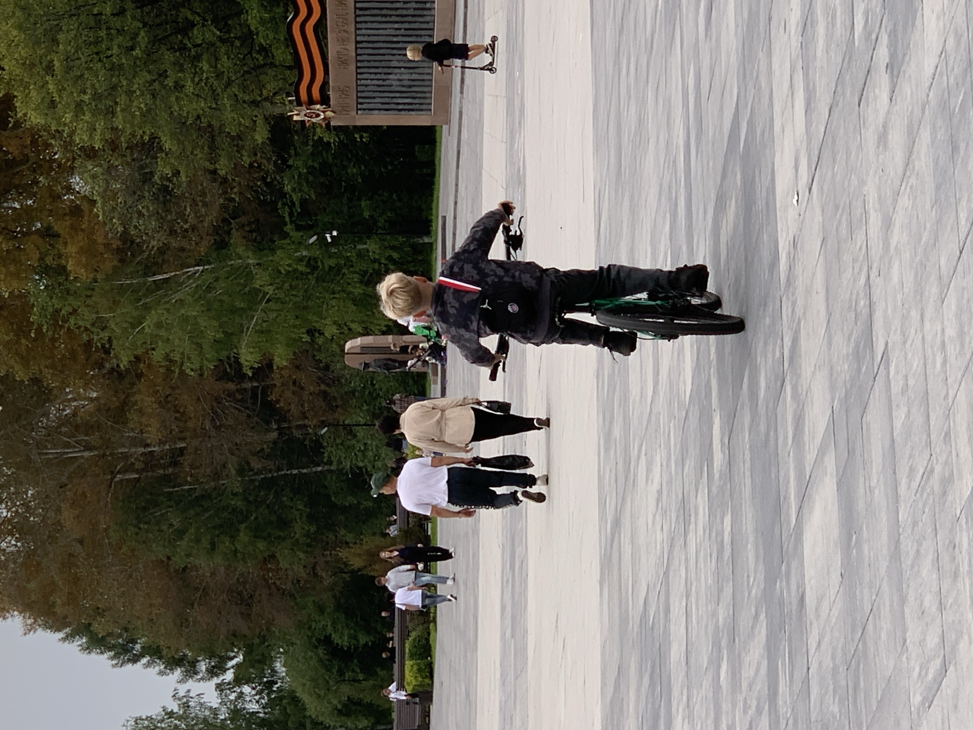 A boy with blond hair is riding a bicycle in a paved outdoor area, seen from behind. He is wearing a dark patterned long-sleeved shirt, dark pants, and black shoes. He has a black backpack with red and white accents. In the background, several people are walking and standing. To the right of the cyclist, another child is riding a scooter. Behind them, a monument with a flag is visible, and trees form a backdrop. The ground is paved with large gray tiles. The lighting suggests an overcast day. The scene appears to be in a park or public square.