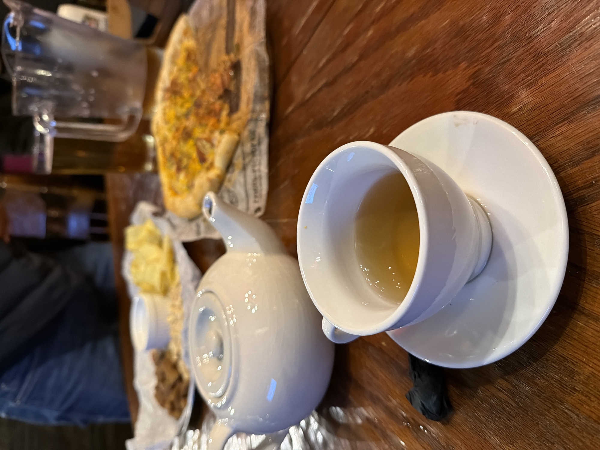 The image shows a table setting with a cup of tea on a saucer next to a teapot, along with some food items including pizza and other dishes. The table appears to be wooden and the overall setting suggests a restaurant or café environment. The location in the city of Chaikovsky, Russia is not discernible from the image.