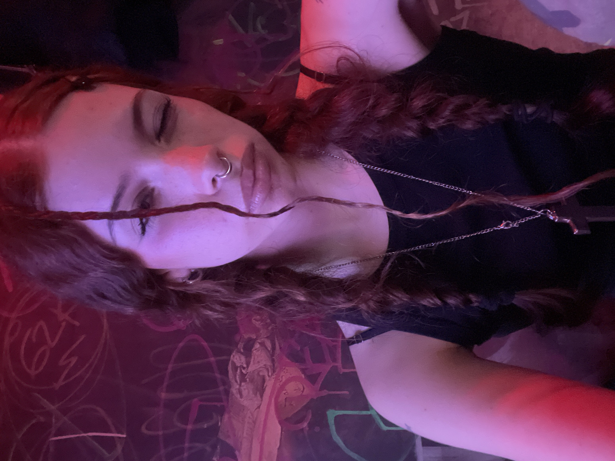 A medium shot depicts a woman with auburn hair in two braids, adorned with a nose ring, wearing a black shirt and a silver cross necklace, eyes closed. She appears to be lying on a surface marked with graffiti in red, white, and green. The scene is illuminated by red and purple light. There is no evidence to suggest this photo was taken in Buenos Aires, Argentina.