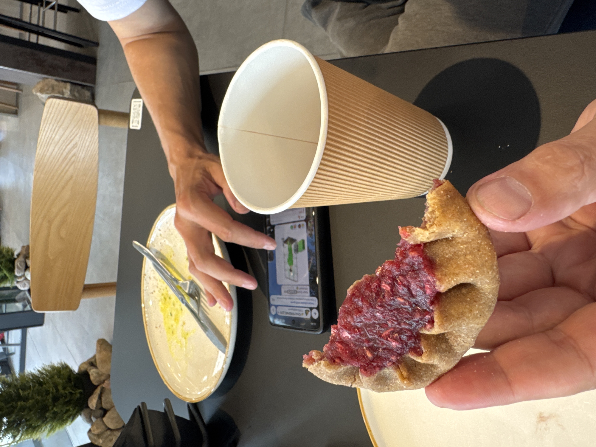 A close-up shot shows a person holding a half-eaten pastry with a red berry filling. The pastry is golden brown with crimped edges and appears to be a type of tart or pie. In the background, a brown corrugated paper cup sits on a black table. To the left of the cup, a smartphone displays a navigation app with a green marker. A fork and knife rest on a white plate with a yellow rim, showing remnants of food. The table is set in what appears to be an outdoor cafe, with a wooden chair and plants visible. The lighting suggests it is daytime. The scene is set in the city of Lakhdenpokhya, Russia.