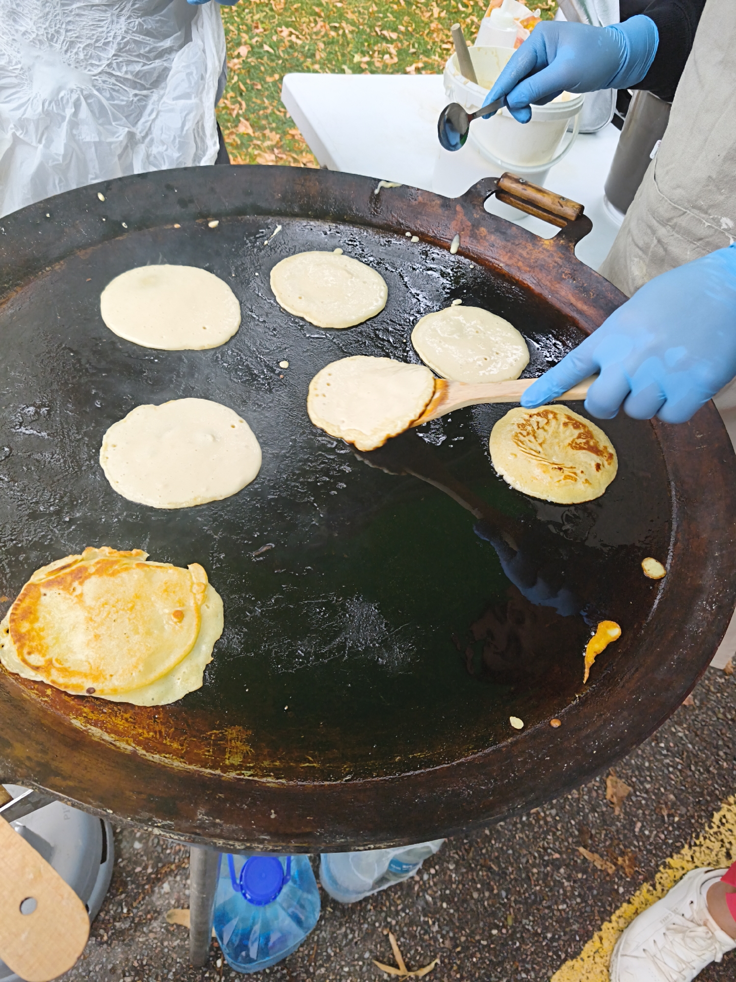 A person wearing blue gloves is cooking pancakes on a large, flat, dark-colored griddle outdoors. The griddle is placed on a stand over a heat source, and there are several pancakes in various stages of cooking. Some are pale yellow, while others are golden brown. A wooden spatula is being used to turn or move a pancake. To the left of the griddle, there is a stack of cooked pancakes. In the background, there is a white table with a bucket and a white container, and some foliage suggesting an outdoor setting, possibly a park or event space. The ground is paved with asphalt and small stones.