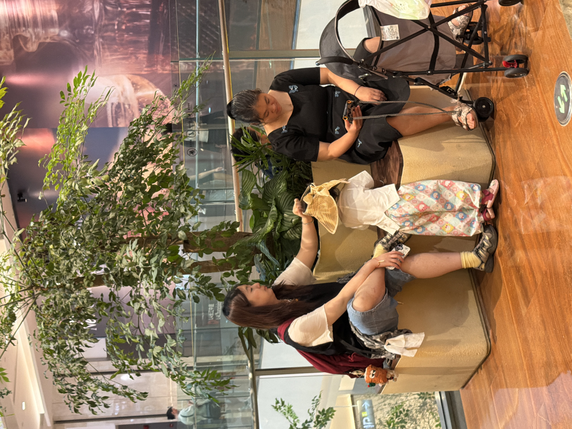 In Yuzhong District, China, two women and a child are seated on a light-colored bench near a large indoor plant. One woman, on the left, wears a white shirt, a black vest, and denim shorts, with a red backpack. She is leaning forward, reaching towards the child. The child, facing away from the camera, is dressed in a white shirt and patterned pants, and is holding a straw hat. The second woman, on the right, is wearing a black dress and is looking down at her phone. Next to her is a dark-colored stroller. The scene appears to be indoors, possibly a shopping mall, with polished wooden flooring and a large mural or advertisement in the background.