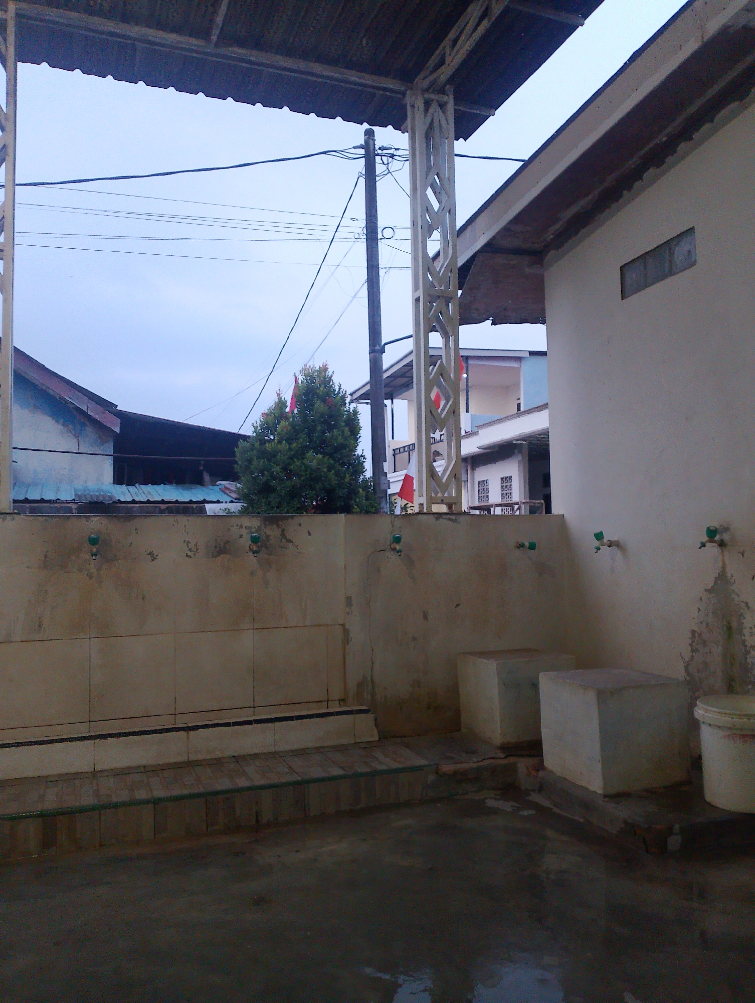 The image depicts an outdoor ablution area, likely for religious purposes, located in Batam, Indonesia. In the foreground, a wet concrete floor is visible, with two cubic concrete structures situated on it. To the right, a white bucket with a red rim is placed. Behind these structures, a beige tiled wall rises, featuring several green faucets attached to it. The tiled wall forms a partition, separating this area from a raised tiled platform with a dark blue trim. A green hose runs along the edge of this platform.
In the middle ground, a dense green tree with a small red flag at its peak stands behind the tiled wall. Further in the background, several buildings are visible. To the left, a light blue and white building with a dark tiled roof is partially obscured. Next to it, a building with a dark, corrugated roof is also present. To the right, a light-colored building with a small rectangular window made of glass blocks is seen. Decorative red flags are also attached to the white support pillars of the structure in the foreground. Overhead, electrical wires are strung across the sky, supported by a utility pole. The sky itself is overcast and pale blue, indicating a possibly dim or early morning light. The overall scene suggests a functional outdoor space, possibly part of a mosque or religious complex.