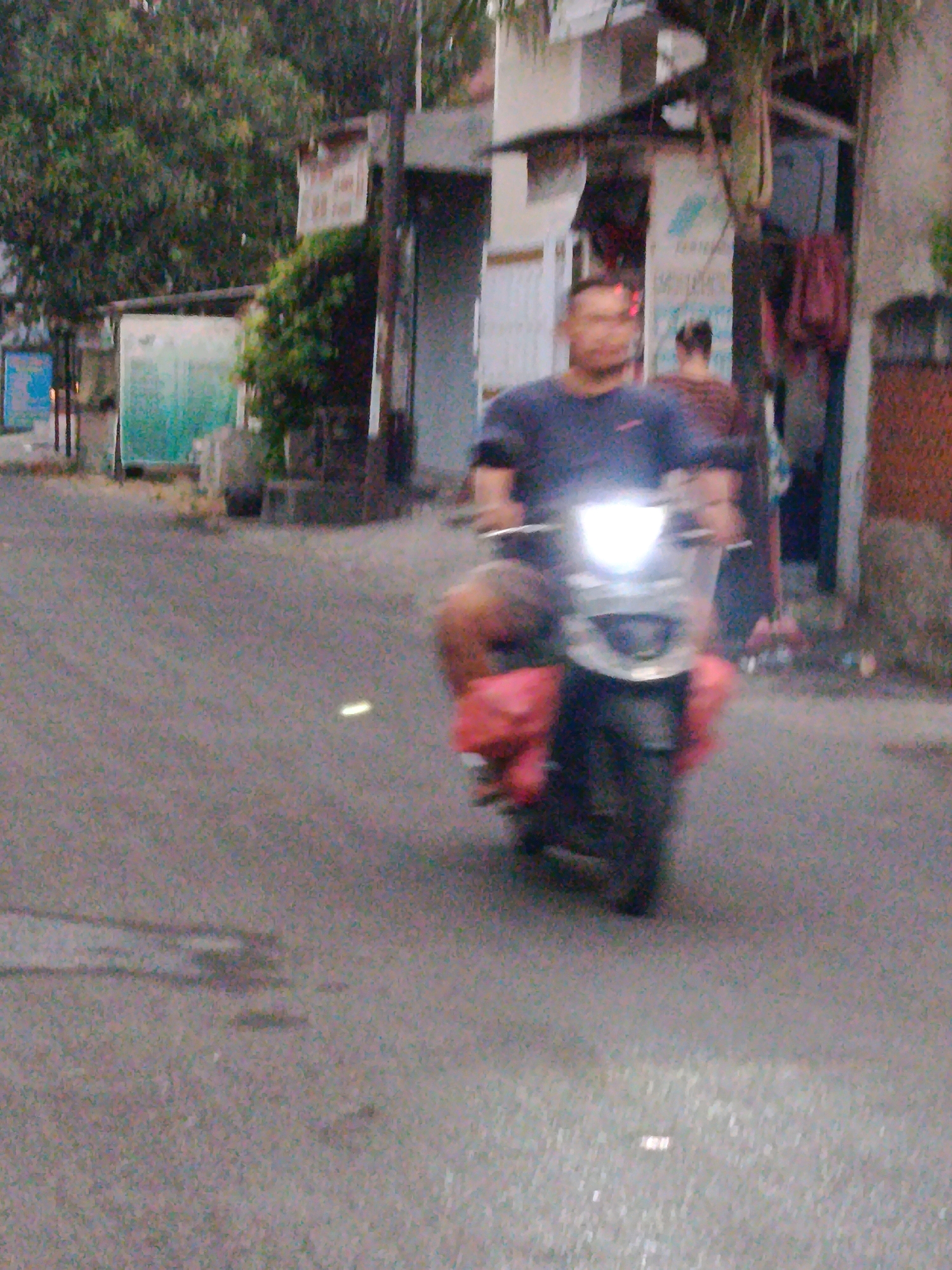 In a city street scene in Bekasi, Indonesia, a man is riding a scooter. He is wearing a dark blue t-shirt and is carrying a bright red bag on the back of his scooter. The scooter's headlight is on, illuminating the road ahead. The road is paved and appears to be wet in places, with some lighter colored patches indicating possibly spilled liquids. On either side of the road, there are buildings with various structures and some vegetation. Trees with dark green foliage are visible in the background, along with what appear to be the facades of shops or houses. The overall lighting suggests it is daytime, possibly overcast or late afternoon, given the muted colors.