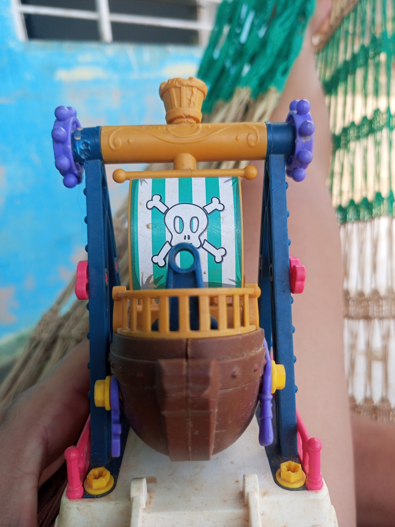 A close-up, eye-level shot captures a miniature toy pirate ship. The ship, predominantly brown, features a yellow deck railing and a white and green striped sail adorned with a skull and crossbones. It is suspended by blue supports connected to an orange overhead bar. Various colorful plastic accents, including purple knobs and pink and yellow levers, are visible on the toy's structure. The toy rests on a white plastic base, possibly part of a larger playset. The background is blurred, with hints of a blue sky with white geometric shapes, and a green and gold patterned fabric or net hanging in the upper right. A person's hand is visible on the left side, holding the toy. The image was taken outdoors, with the sunlight casting shadows and highlights on the toy. The location in La Tagua, Venezuela, is not discernable from the visual information provided.