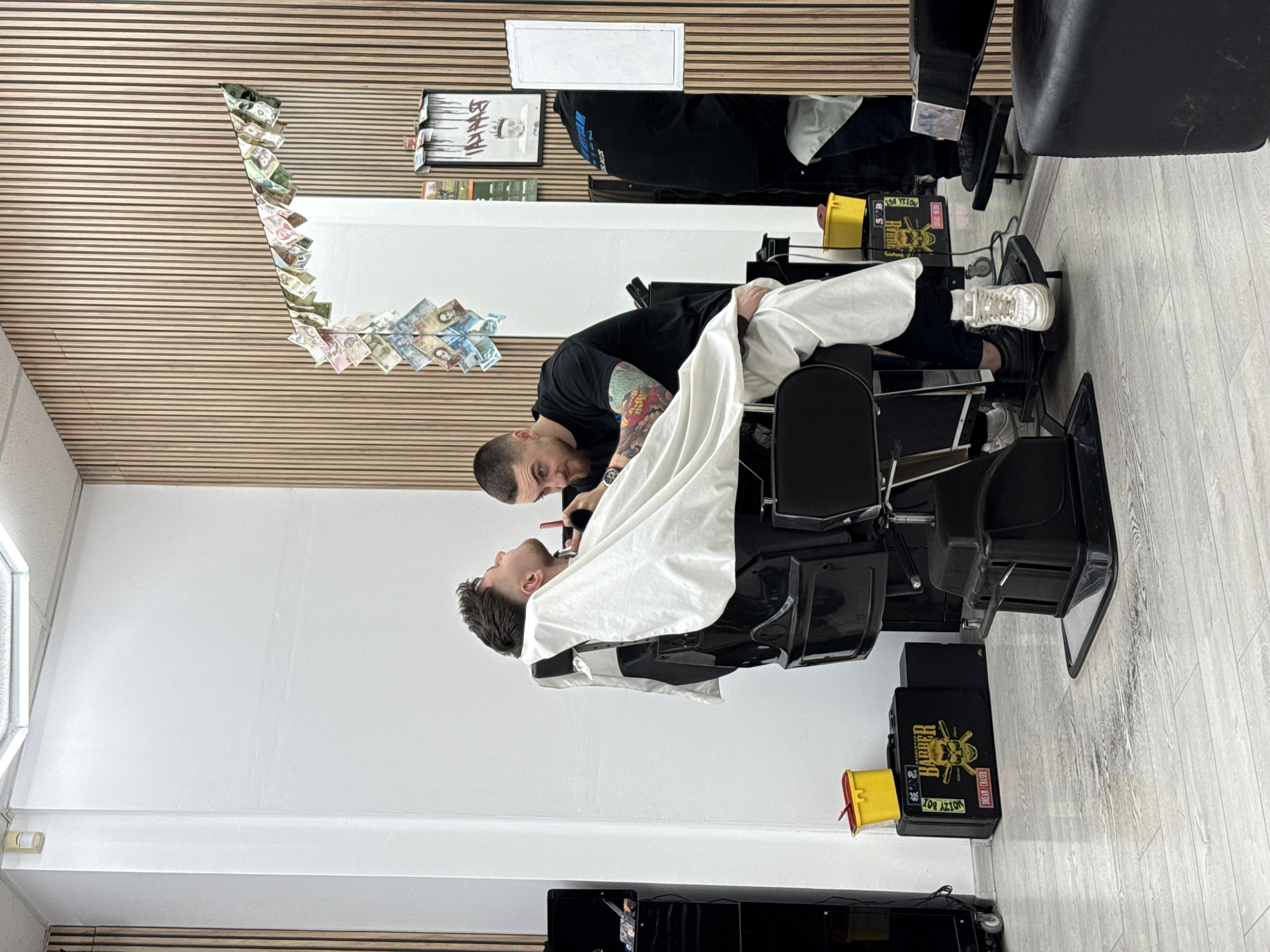 A barber is giving a shave to a customer in a barber shop. The barber is standing next to the customer, who is lying back in a barber's chair with a white cloth over his torso. The barber is using a straight razor. The barber has a sleeve tattoo on his right arm. There are various items on the wall, including money, a framed picture, and a mirror. The shop is located in Sofia, Bulgaria, based on the presence of Bulgarian currency adorning the mirror.