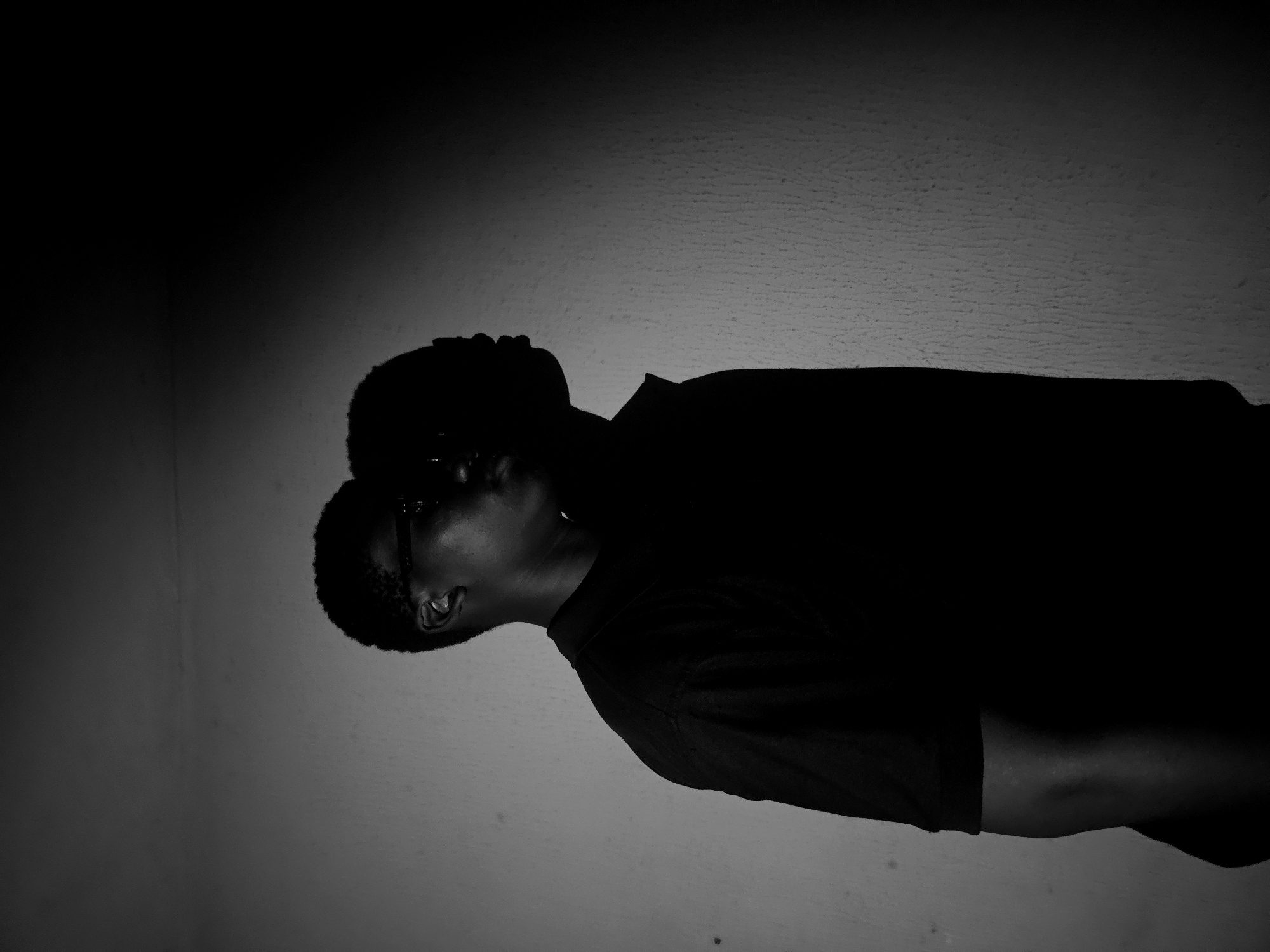 The image is a black and white photograph showing a person. The person is wearing glasses and a dark shirt. The environment appears to be indoors against a wall. The location is Unknown, Unknown.