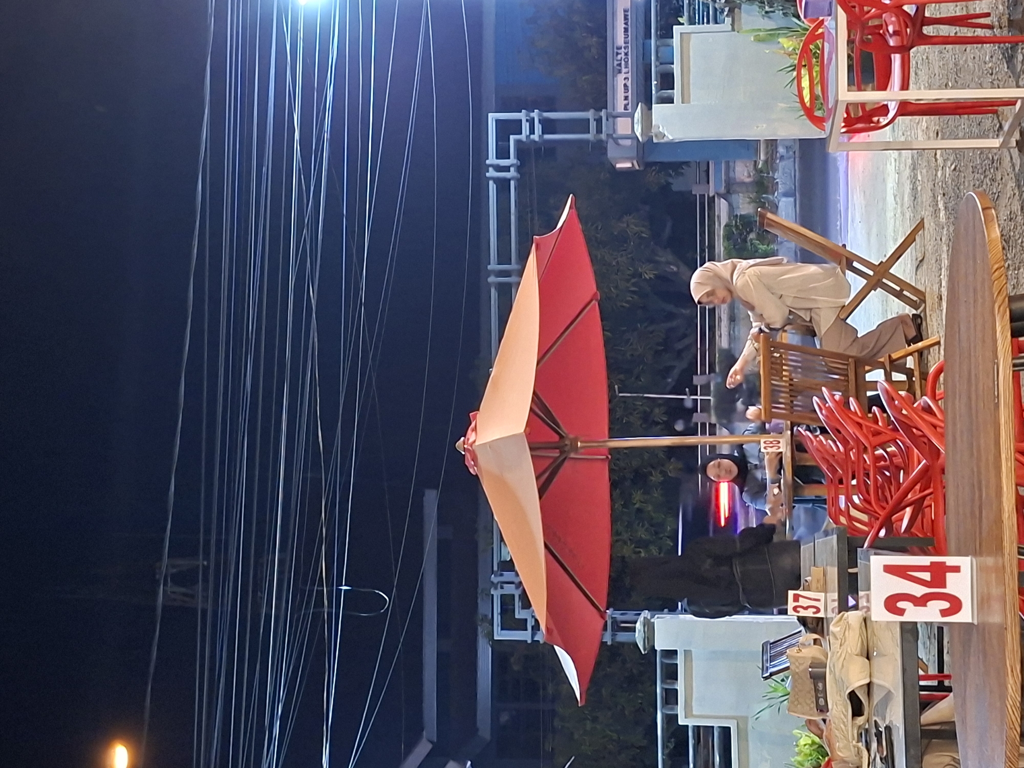 The image shows an outdoor scene, possibly a restaurant or café, at night in the city of Lhokseumawe, Indonesia. A tall, narrow umbrella with red and beige panels is a central feature. Numerous thin cables or wires are visible against the dark sky, seemingly related to utility infrastructure. In the background, a person wearing a headscarf and light-colored clothing is interacting with a wooden structure, possibly rearranging chairs. Several red chairs are stacked nearby. Table numbers "34" and "37" are prominently displayed on the tables. Other people are present in the midground, including one wearing a dark jacket. A sign partially visible in the background reads "VALTE PLN UP3 LHOKSEUMAWE". The lighting suggests an urban environment at night, likely captured in a public space.
