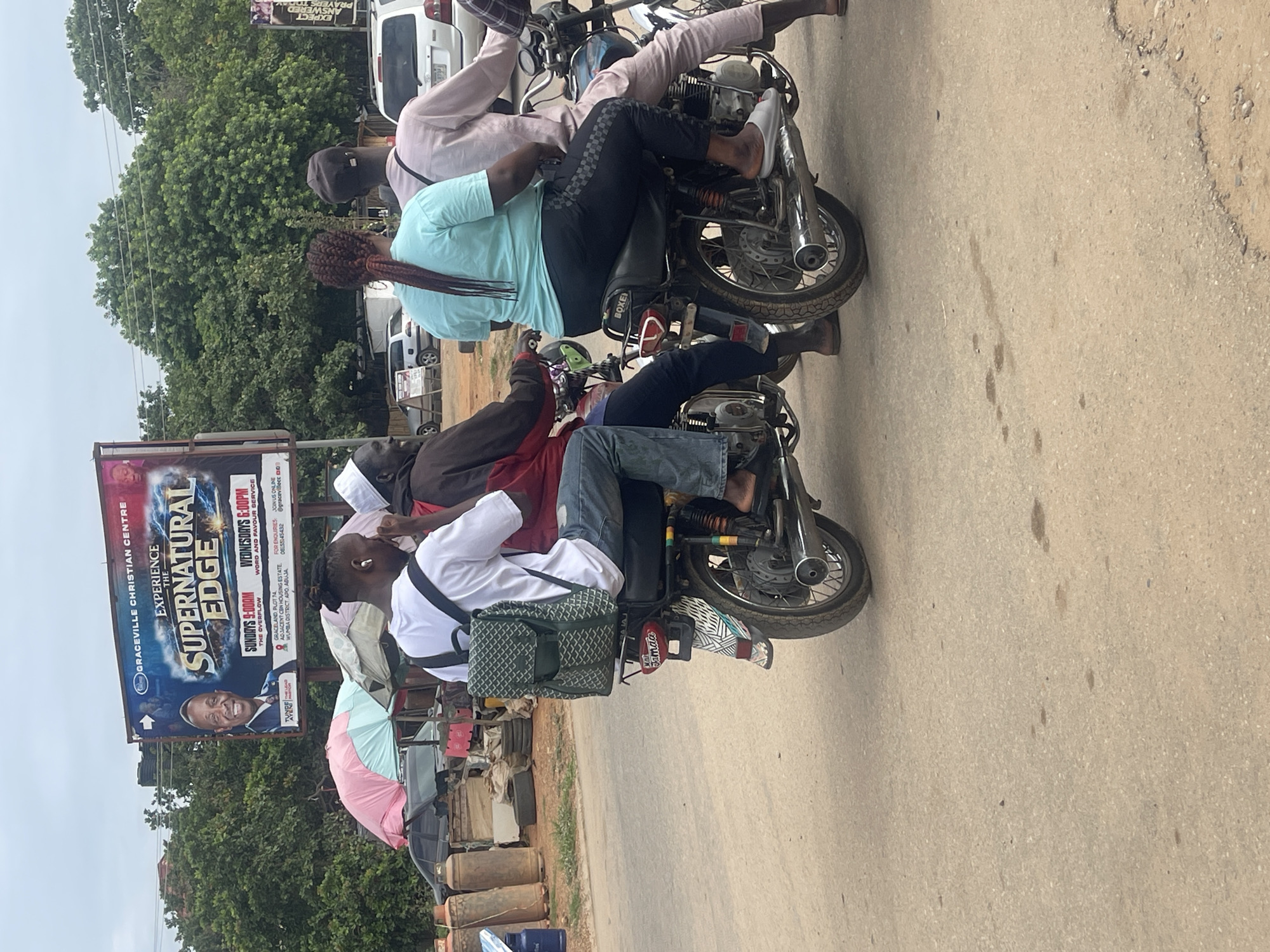 The image depicts a street scene in Abuja, Nigeria, featuring several individuals on motorcycles. In the foreground, a person wearing a white t-shirt and jeans rides a motorcycle, carrying a green patterned backpack. Behind them, another person on a motorcycle wears a light blue t-shirt and black pants with a checkered pattern. To the right, a third individual in a pink outfit is also on a motorcycle. The background includes a billboard for "GRACEVILLE CHRISTIAN CENTRE EXPERIENCE THE SUPERNATURAL EDGE" with event details for Sundays and Wednesdays. Trees and other vehicles are visible in the distance. The overall environment appears to be an urban street with moderate traffic.