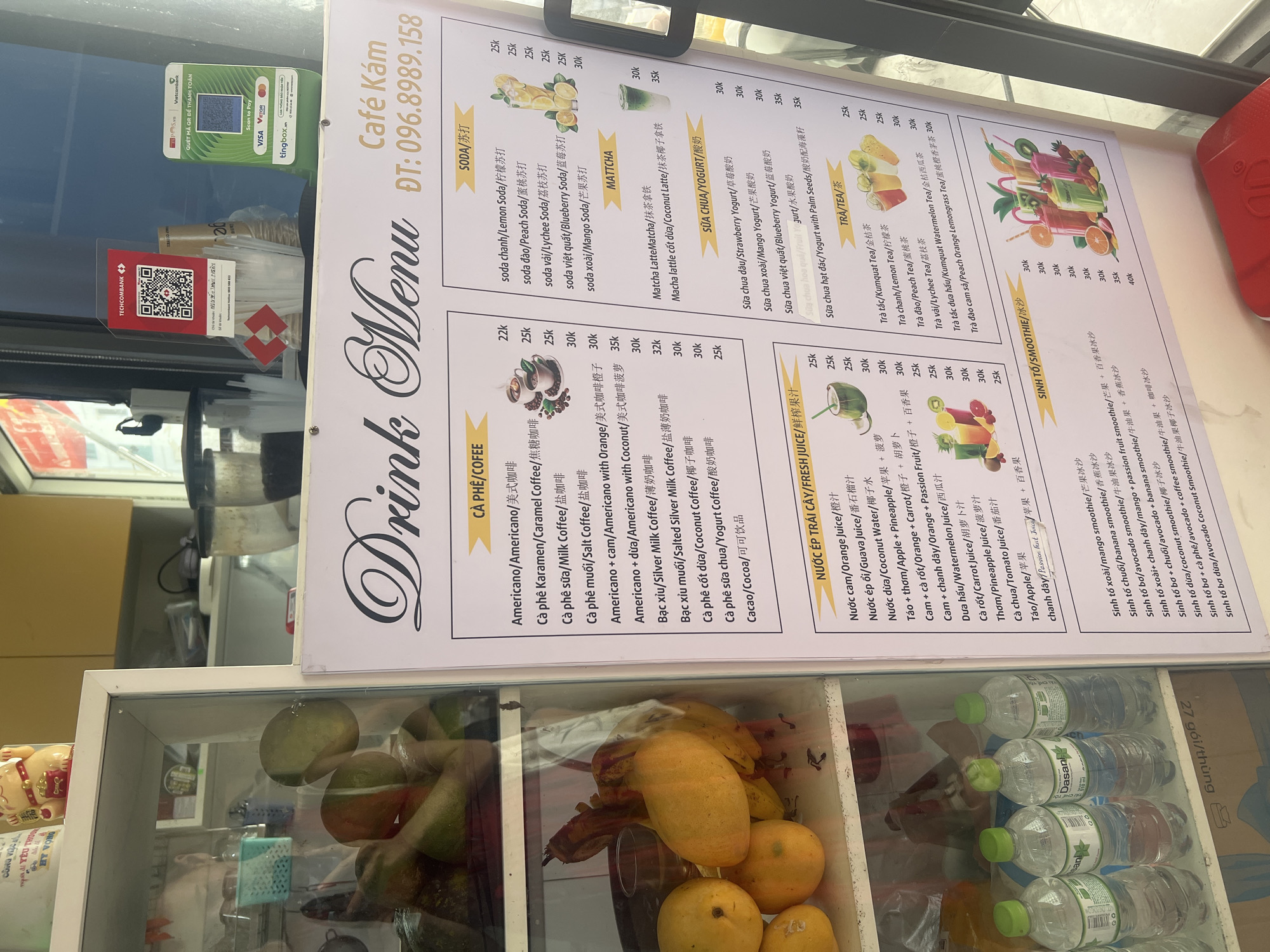 The image captures a detailed view of a drink stall's menu and display counter, likely located in Bắc Nha Trang, Vietnam, as indicated by the context. The main subject is a large, brightly lit menu board titled "Drink Menu" from "Café Kám," with a contact number "ĐT: 096.89.158".
The menu is presented in Vietnamese, English, and Chinese, organized into several categories: CÀ PHÊ/COFFEE, SODA/苏打, MATTCHA, SỮA CHUA/YOGURT/酸奶, NƯỚC ÉP TRÁI CÂY/FRESH JUICE/鲜榨果汁, TRÀ/TEA/茶, and SINH TỐ/SMOOTHIE/冰沙. Each item has a corresponding price listed in "k," signifying Vietnamese Dong (e.g., Americano for 22k, Lemon Soda for 25k, Matcha Latte for 30k, Strawberry Yogurt for 30k, Orange Juice for 25k, Avocado Smoothie for 35k).
To the left of the menu, two QR code payment stands are visible. One prominently displays "Vietcombank" and "QUÉT MÃ QR ĐỂ THANH TOÁN" (Scan QR code to pay), also mentioning "Scan to Pay" with logos for Visa, VietQR, and Tingbox.vn. The second stand is for "TECHCOMBANK" with a QR code and account name "NGUYỄN THU NGÂN".
Below the menu and payment stands, a glass display counter holds various fresh fruits, including ripe bananas, mangoes, and what appear to be guavas or avocados, alongside multiple bottles of Dasani water, some of which are labeled "27 gói/thùng" (27 packs/carton). A golden lucky cat figurine is also visible on the left side of the counter. The overall scene suggests a vibrant, modern establishment catering to a diverse clientele.