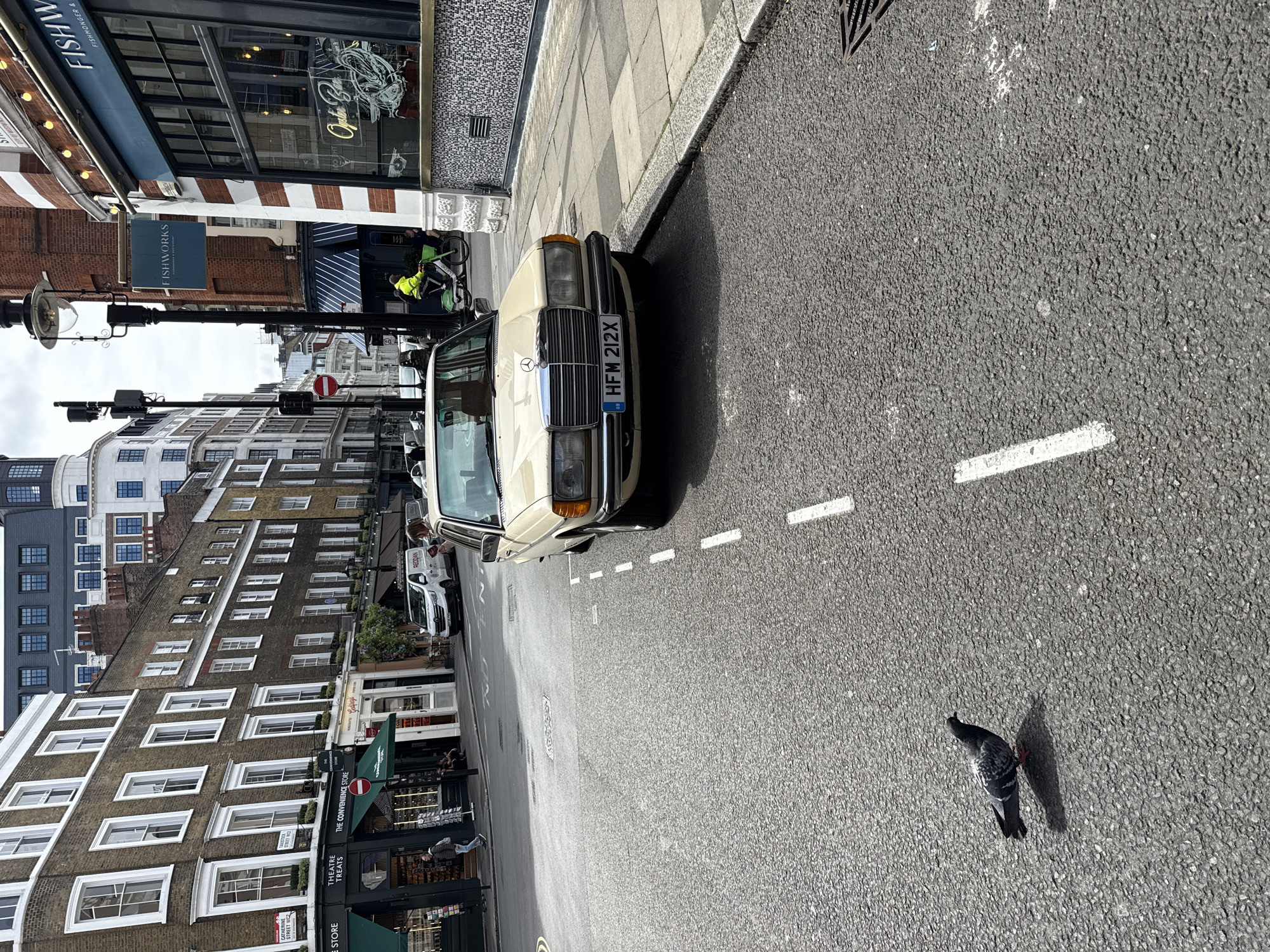 The image shows a street scene in London, United Kingdom. A beige Mercedes-Benz with the license plate "HFM 212X" is parked along the right side of the street. A pigeon stands on the asphalt road surface in the lower right corner. The street has a white dashed line indicating lanes. Buildings line the left side of the street, including businesses like "THEATRE TREATS," "THE CONVENIENCE STORE," and "FISHWORKS." Street signs indicate "CATHERINE STREET WC2". A delivery cyclist wearing a yellow jacket is near the Fishworks restaurant. The sky is overcast.