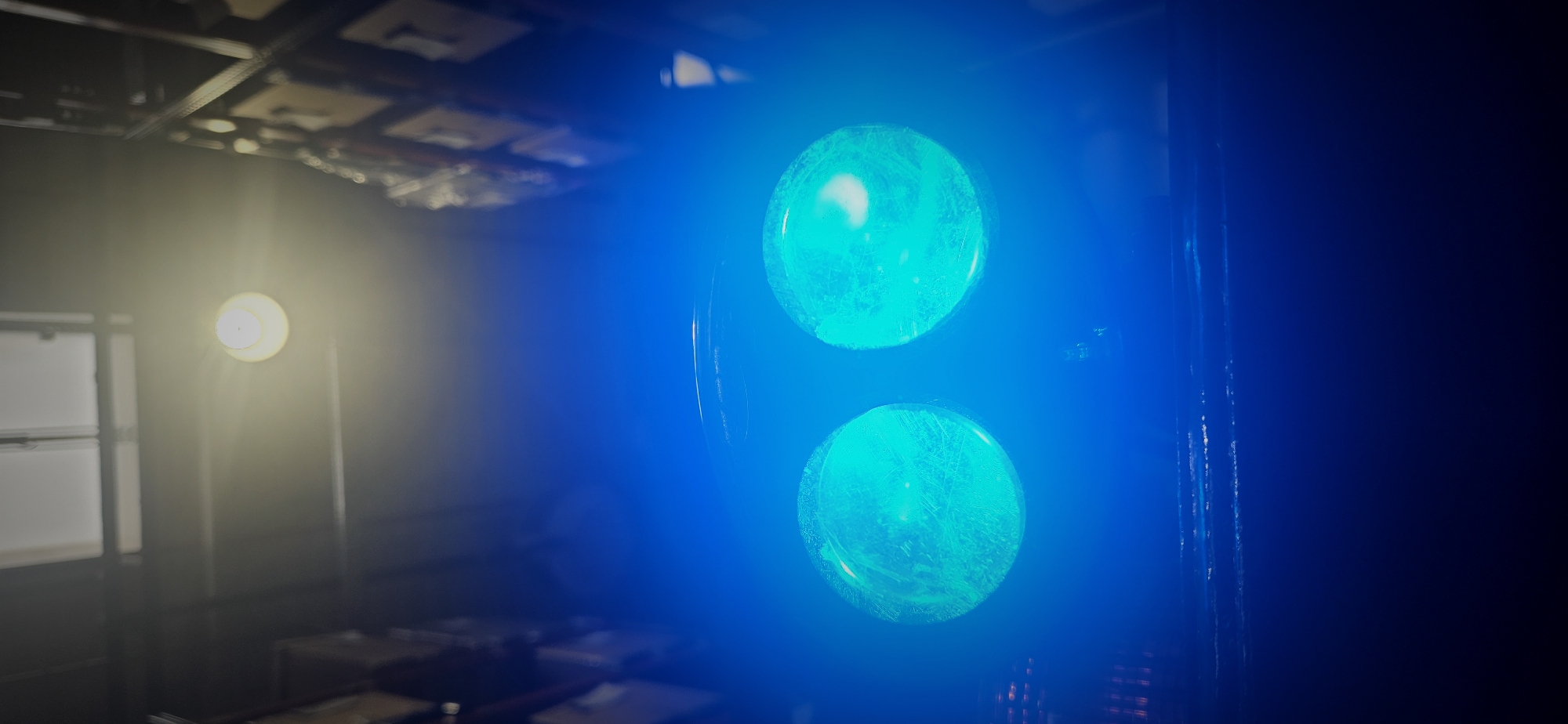 The image displays an indoor scene, likely a dimly lit room or specialized environment, with prominent artificial lighting. On the right side of the frame, two large, circular lights emit a strong, vibrant blue glow. Their surfaces appear textured or frosted, with some lighter internal reflections. These blue lights seem to be part of a larger fixture or piece of equipment, possibly a control panel, an indicator, or stage lighting.
To the left, further into the background, a single circular light source emits a warm, yellow-white glow, illuminating a portion of the space with a softer light. The intense illumination from both the blue and yellow lights creates lens flares and a hazy atmospheric effect around them.
The background elements are mostly dark and out of focus, suggesting a shallow depth of field focusing on the lights. In the upper left, a ceiling with square panels is visible, hinting at an architectural setting like an office, studio, or industrial space. Indistinct, blurry shapes can be seen in the lower left, possibly furniture or other equipment. The lighting conditions suggest that these artificial lights are the primary light sources in an otherwise dark setting. No people or visible text are present in the image. The location context for this media file is Plovdiv, Bulgaria.