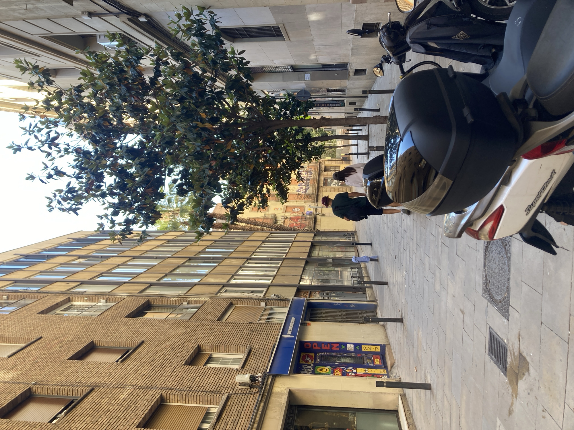 The image captures an urban street scene in Barcelona, Spain. A multi-story building with a brick facade and numerous windows dominates the left side of the frame. At street level, a shop with a blue awning is visible, displaying a sign that reads "OPEN." A tree with dense foliage partially obscures the view of the building and street. In the background, graffiti decorates the walls of buildings further down the street. Two people are walking on the sidewalk. A parked scooter occupies the right side of the image, taking up a significant portion of the frame. The sidewalk is paved with light-colored stone tiles. The scene is bathed in natural light, creating a sunny atmosphere. The overall impression is of a typical urban environment in Barcelona.