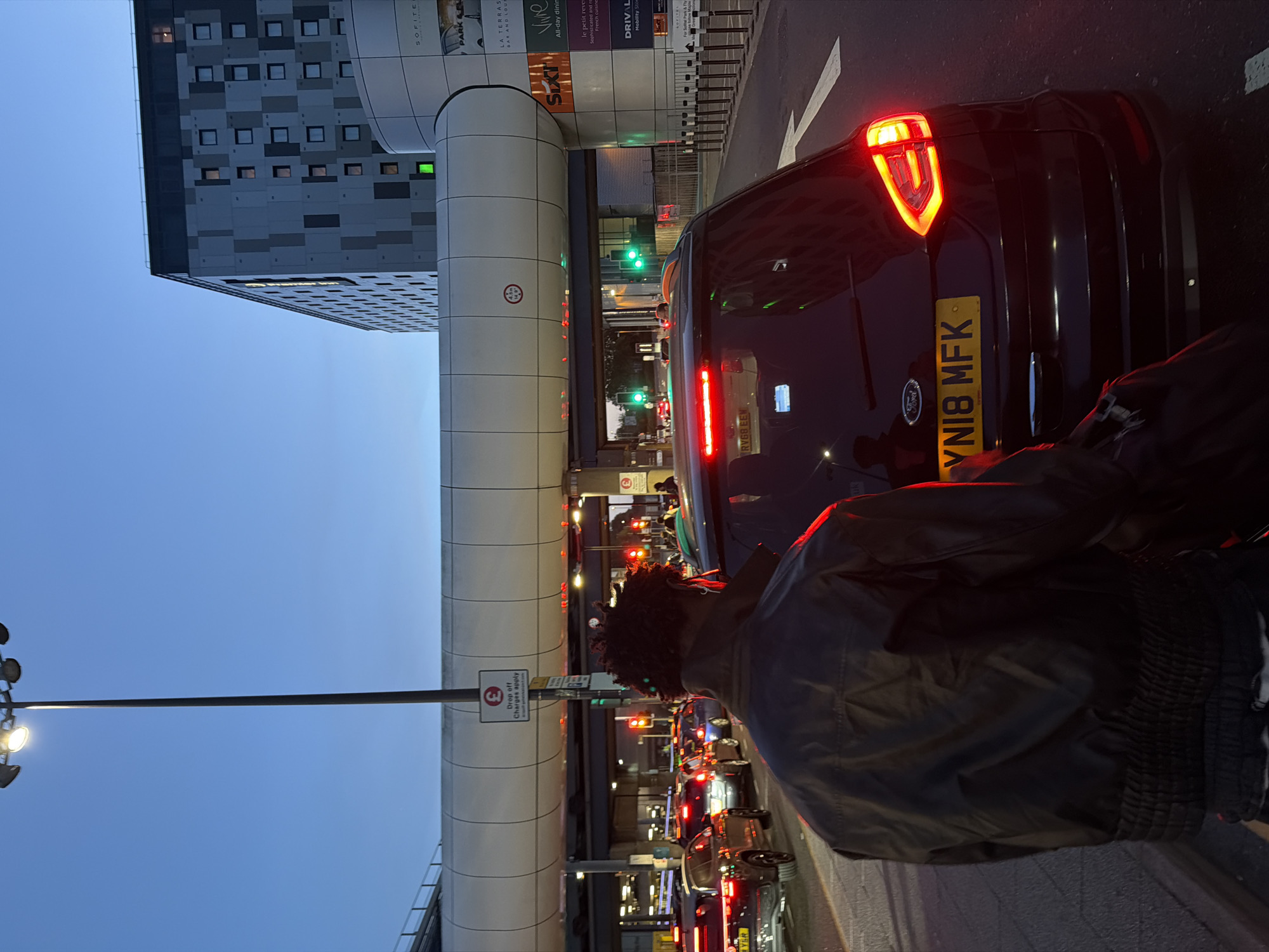 The image depicts a street scene at dusk, likely in Crawley, United Kingdom, given the presence of a UK license plate and what appears to be a sign for Heathrow Airport. In the foreground, a person with dark, curly hair is seen from the back, wearing a black jacket. They are standing on a paved area, possibly a sidewalk or street, next to a black SUV.
The SUV has its rear brake lights illuminated, and a yellow UK license plate reading "YN18 MFK" is visible. The vehicle is stopped in traffic, indicated by the presence of other vehicles in the background with illuminated brake lights. Traffic lights are visible, some showing red, others green, suggesting an intersection or traffic control point.
Above the road, a modern, elevated walkway or bridge structure spans across the scene. This structure has a light-colored, rounded facade with various signs attached to its side. One sign displays a pound sterling symbol and the text "Drop off Charges Apply". Another sign indicates a height restriction of "4.5m (14'9")". In the background, a large, modern building with a patterned facade and numerous windows rises against the twilight sky. The overall atmosphere is that of a busy urban environment during evening traffic.