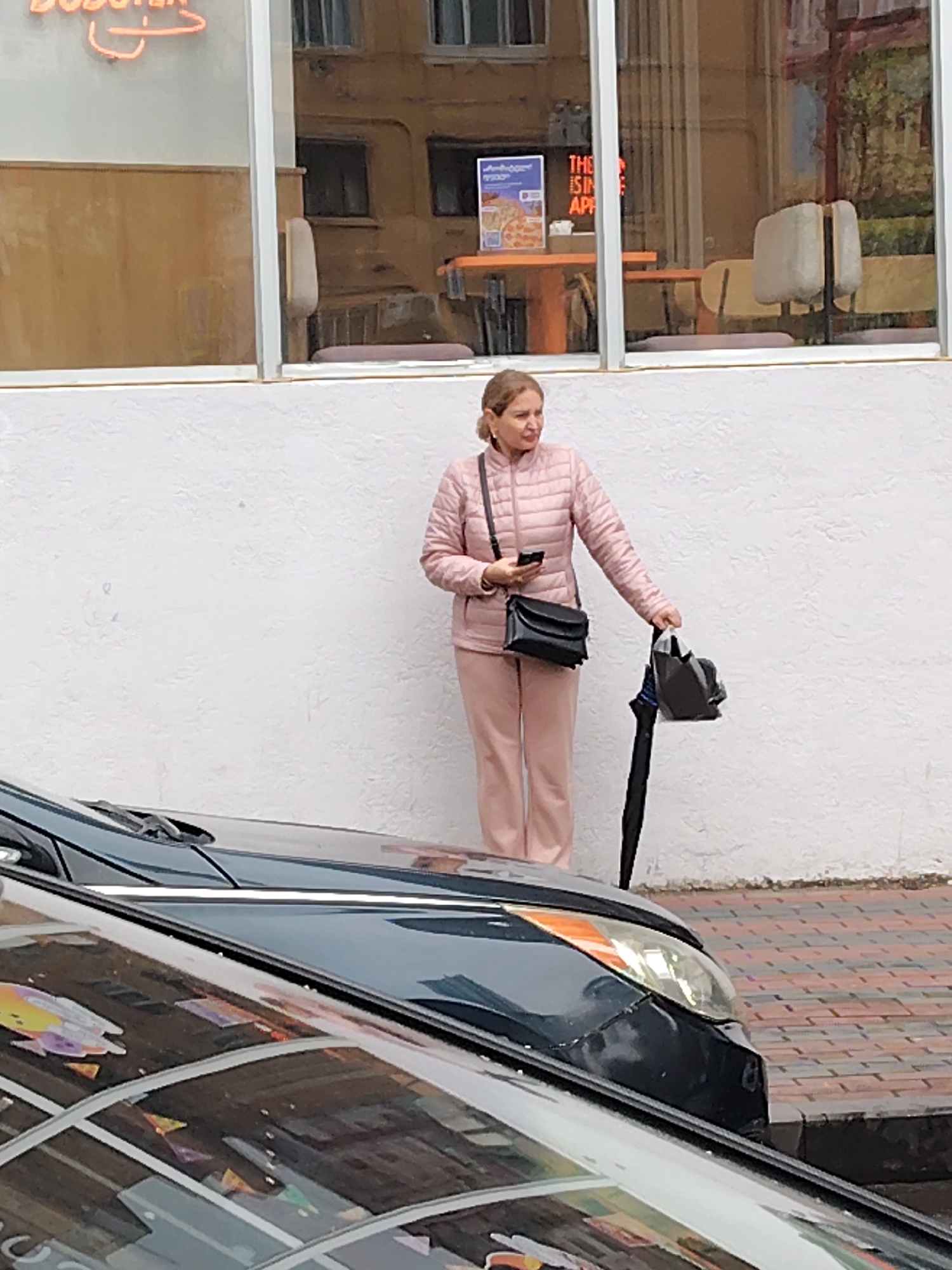 A woman stands on a brick-paved sidewalk in Batumi, Georgia. She wears a light pink quilted jacket and matching pants. A black purse hangs across her shoulder, and she holds a smartphone in one hand. In her other hand, she holds a black umbrella and a dark gray bag. A black car is parked in front of her, partially obscuring the view. Behind her, a white wall is visible with a large window reflecting a building with orange accents. A sign with neon lettering is above the window, and a poster is visible inside the window.