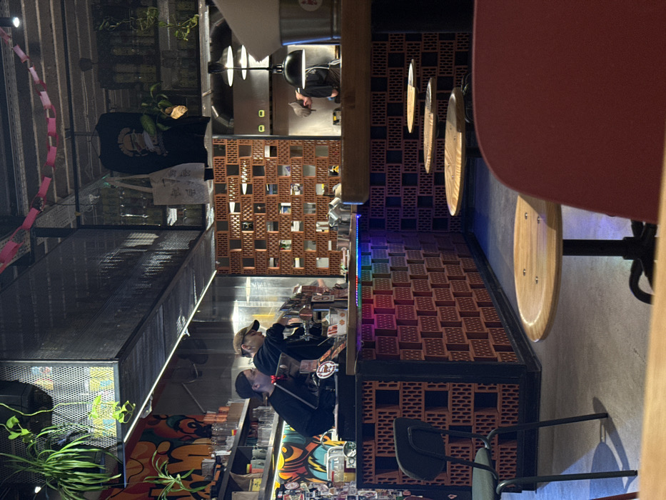 A modern interior features a bar constructed with red brick-like blocks, interspersed with square cutouts, and illuminated by strip lighting. Adjacent to the bar, round wooden tables with metal chairs are visible. Patrons are seated at the bar, while a chef is positioned further back. Hanging circular decorative elements add a stylish touch to the space, and plants are placed near the entrance, contributing to the overall ambiance.
