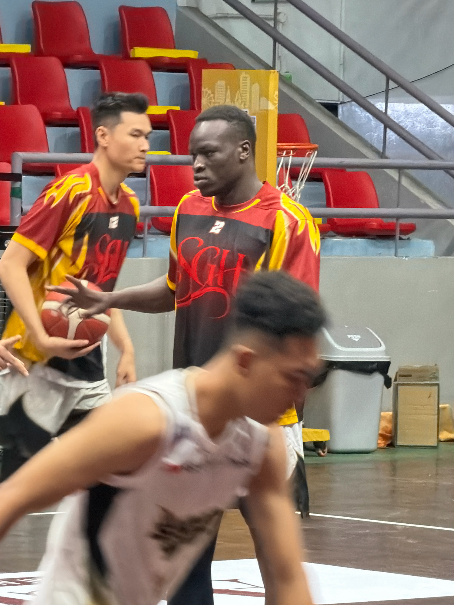 The image depicts a basketball game in progress on an indoor court. In the foreground, a player in a white jersey is blurred, with their arm extended and body in motion. Behind them, two players are more clearly visible. One player, wearing a red and yellow jersey with "SGH" emblazoned on it, is holding a basketball and looking towards the left. Another player, also in a similar red and yellow jersey, stands to his right, also looking towards the left. The background features red stadium seating, a trash can, and a yellow billboard with a graphic. The scene is likely within a sports facility. There is no specific indication of the location being Nha Trang, Vietnam.
