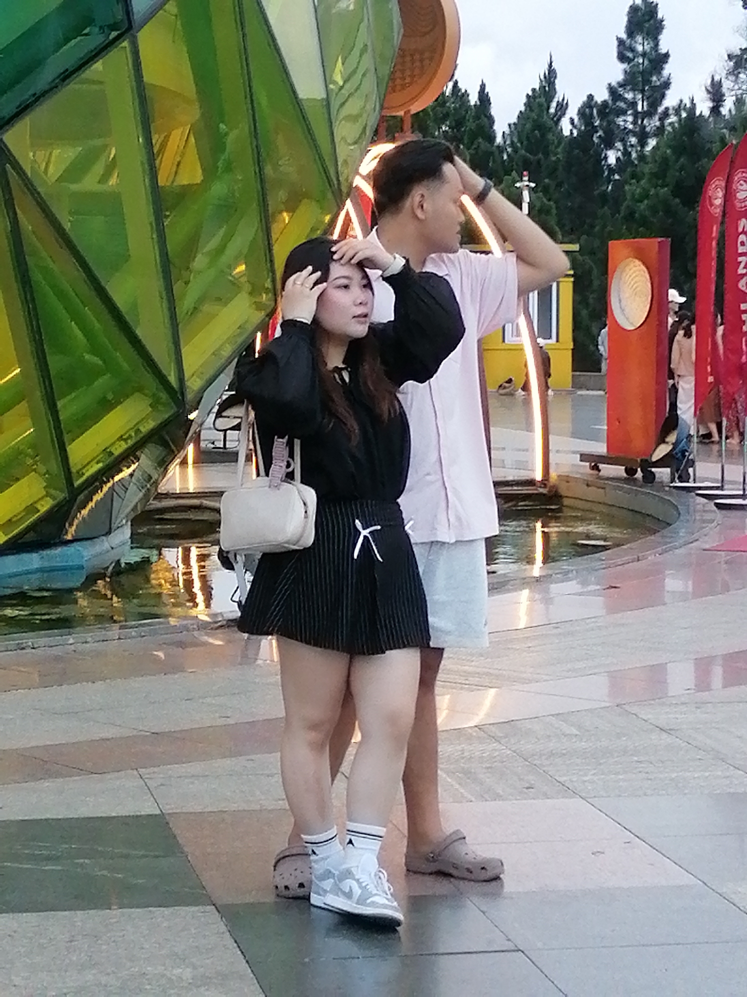 Two individuals, a woman and a man, stand in front of a large, angular, green, faceted sculpture with yellow and green colored glass panels. The woman, on the left, wears a black long-sleeved top with puffy sleeves, a black pleated skirt with white pinstripes, white socks, and light grey sneakers. She carries a small, light-colored crossbody bag and has her hands near her head. The man, on the right, wears a pale pink short-sleeved collared shirt and light grey shorts. He has one hand raised to his head. They appear to be posing or looking around. In the background, there is a fountain with water, a yellow structure, red flags, and trees. The ground is paved with square tiles in shades of beige, grey, and green. The overall scene suggests an outdoor public space, possibly a park or plaza in Da Lat, Vietnam, given the presence of the distinctive architecture and foliage.