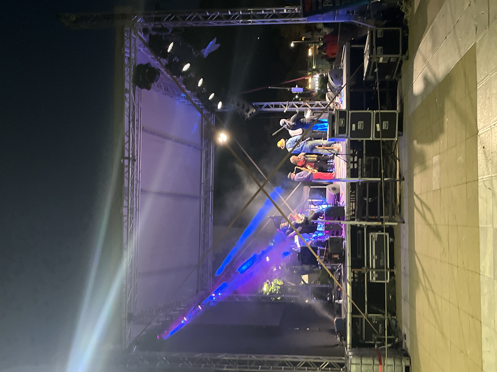 An outdoor nighttime concert stage is set up with a band performing. The stage is lit by a mix of purple and blue lights, with spotlights creating beams that cut through the smoke or haze in the air. Several musicians are visible on stage, some playing instruments like a saxophone, guitar, and drums. Stage equipment, including speakers and cases, is stacked on and around the stage. In the background, there are some lights from buildings and trees, suggesting an urban or town setting. A crowd of people is partially visible in the distance, seated or standing. The foreground shows a paved area, possibly a walkway or plaza.