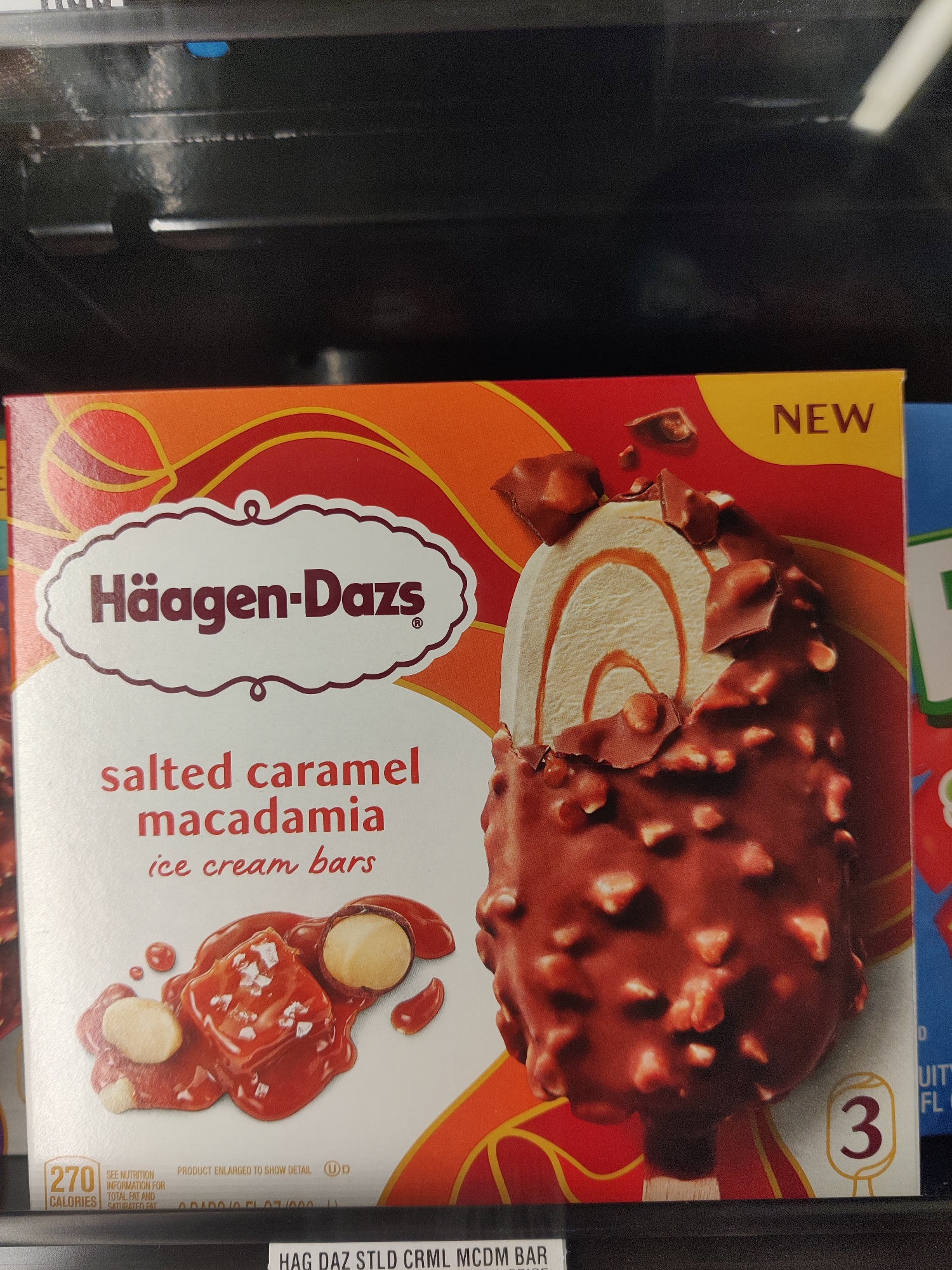 The image shows a close-up of a box of Häagen-Dazs Salted Caramel Macadamia ice cream bars. The box features a prominent image of a chocolate-covered ice cream bar with caramel swirls and macadamia nuts. The text on the box indicates the flavor and brand. The lower portion of the box displays nutritional information and the number "3", likely indicating the quantity of bars in the package. A black shelf or display case is visible at the bottom of the image. The background is dark and out of focus, suggesting it is inside a store or freezer. The image was taken in Mechanicsburg, United States of America.