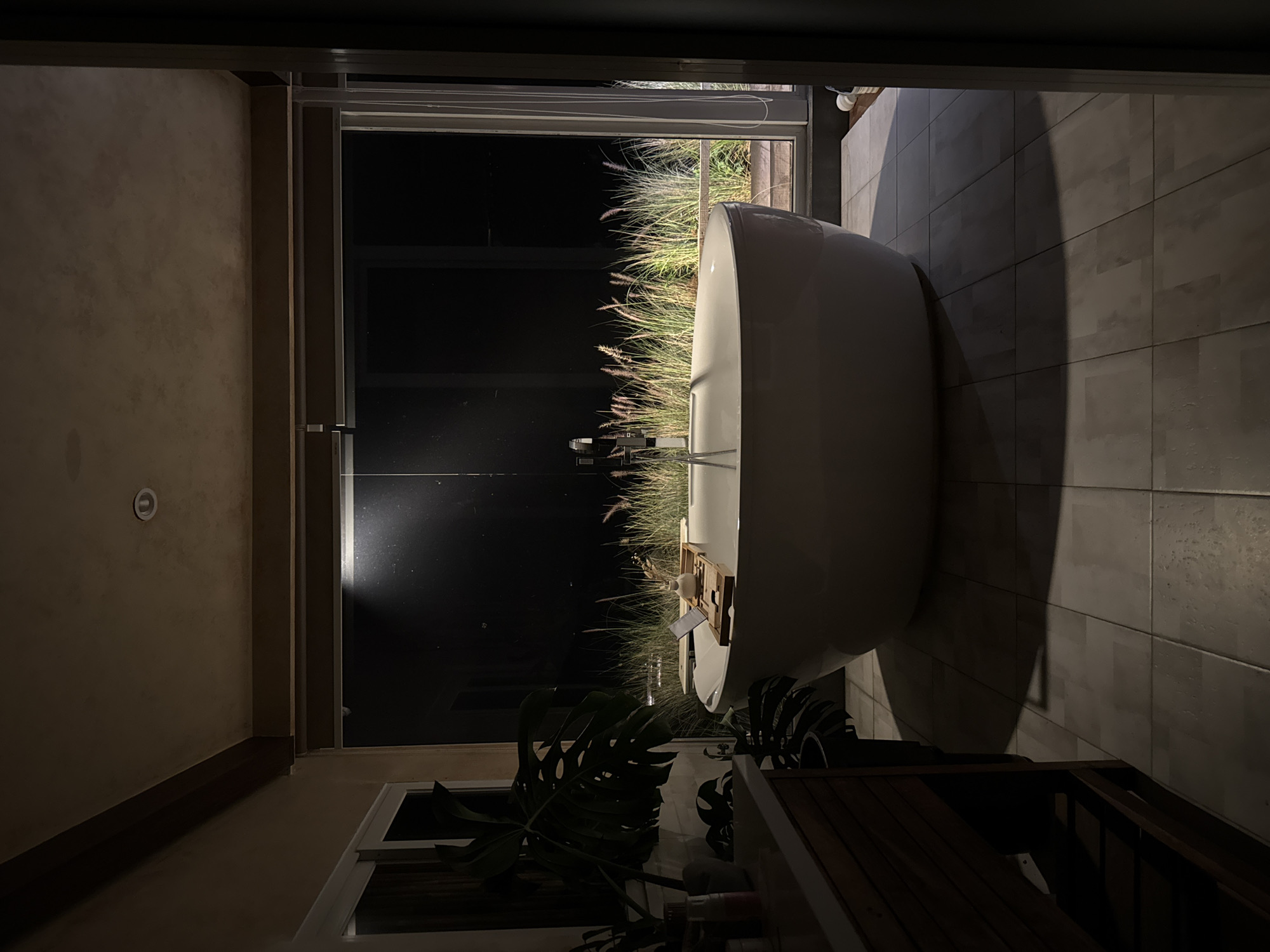The image depicts a modern bathroom with a large, freestanding bathtub as the central focus. The bathroom features a tiled floor and a large glass window that looks out onto a dimly lit outdoor area with tall grasses illuminated by spotlights. There is a wooden bath tray across the tub, holding a small vase with dried flowers and what appears to be a laptop. To the left of the tub is a wooden vanity with drawers, and a large potted plant with broad leaves is visible in the foreground. The overall ambiance is dark and tranquil, suggesting it is nighttime. The image does not provide any specific visual cues to pinpoint its location in Da Lat, Vietnam.