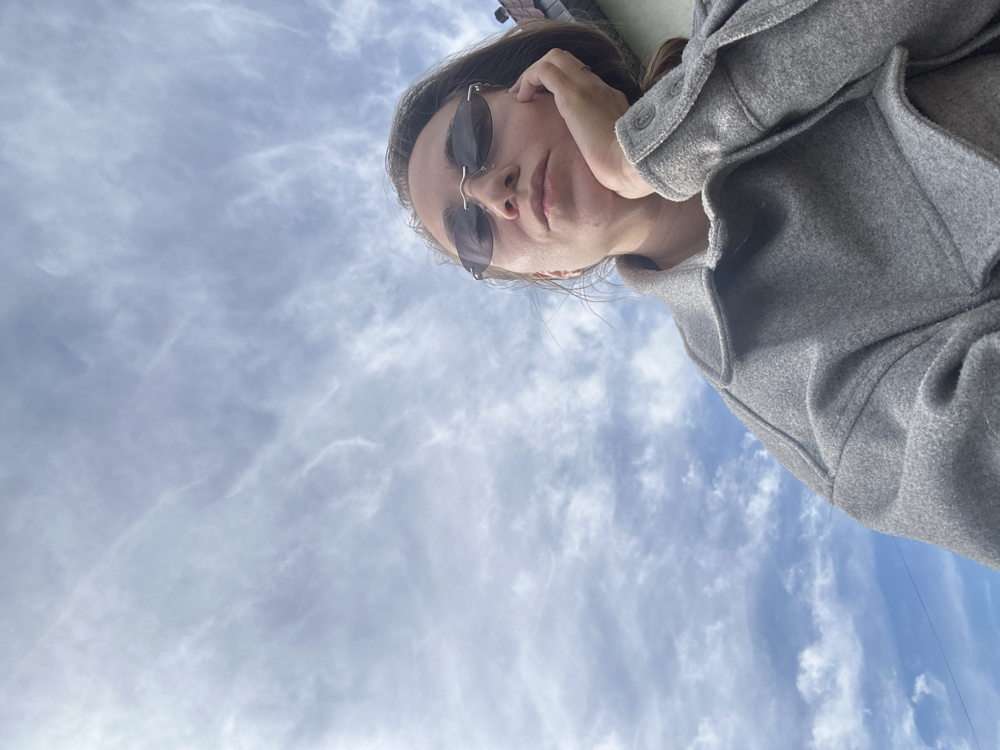 The image is a unique low-angle shot, looking upwards, capturing a young woman against a vast expanse of sky in Kharnavy, Russia. The woman is positioned diagonally in the upper right portion of the frame, her head angled towards the bottom left. She has medium brown hair and is wearing small, oval-shaped, dark-lensed sunglasses. Her right hand rests gently on her cheek or jawline, suggesting a thoughtful or relaxed pose. Her expression is neutral and calm.
She is dressed in a grey, textured, long-sleeved jacket or coat. Details like visible buttons on the left sleeve cuff and deep front pockets are discernible. The sky dominates the majority of the image, showcasing a partly cloudy daytime scene. Wispy, streaky white clouds are scattered across a pale blue background, with some denser cloud formations also present. The weather appears fair and bright. No specific architectural or landscape features are visible to denote a precise location within Kharnavy, Russia, beyond the sky itself. No text is visible in the image.