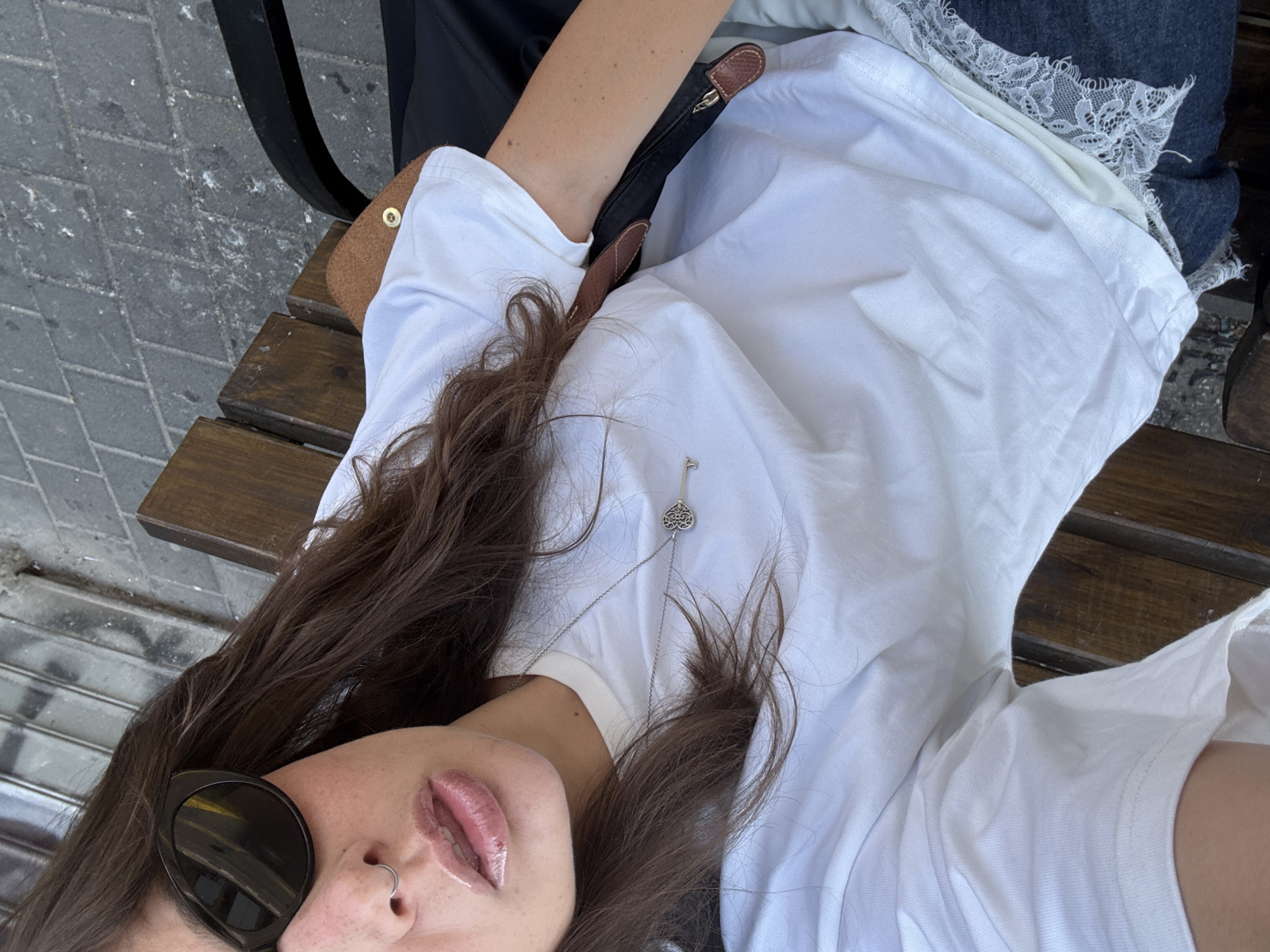 A female subject is depicted in a selfie taken outdoors. She is wearing a white t-shirt and a silver key necklace. She has brown hair and is wearing sunglasses and a nose ring. She is seated on a wooden bench, which appears to be located in a city setting, indicated by the gray, brick-paved ground visible at the upper left. Her shoulder bag sits next to her on the bench. The presence of a bench suggests an urban environment. Further details, such as street signs, architecture, or local flora, are required to confirm the Tel-Aviv location.