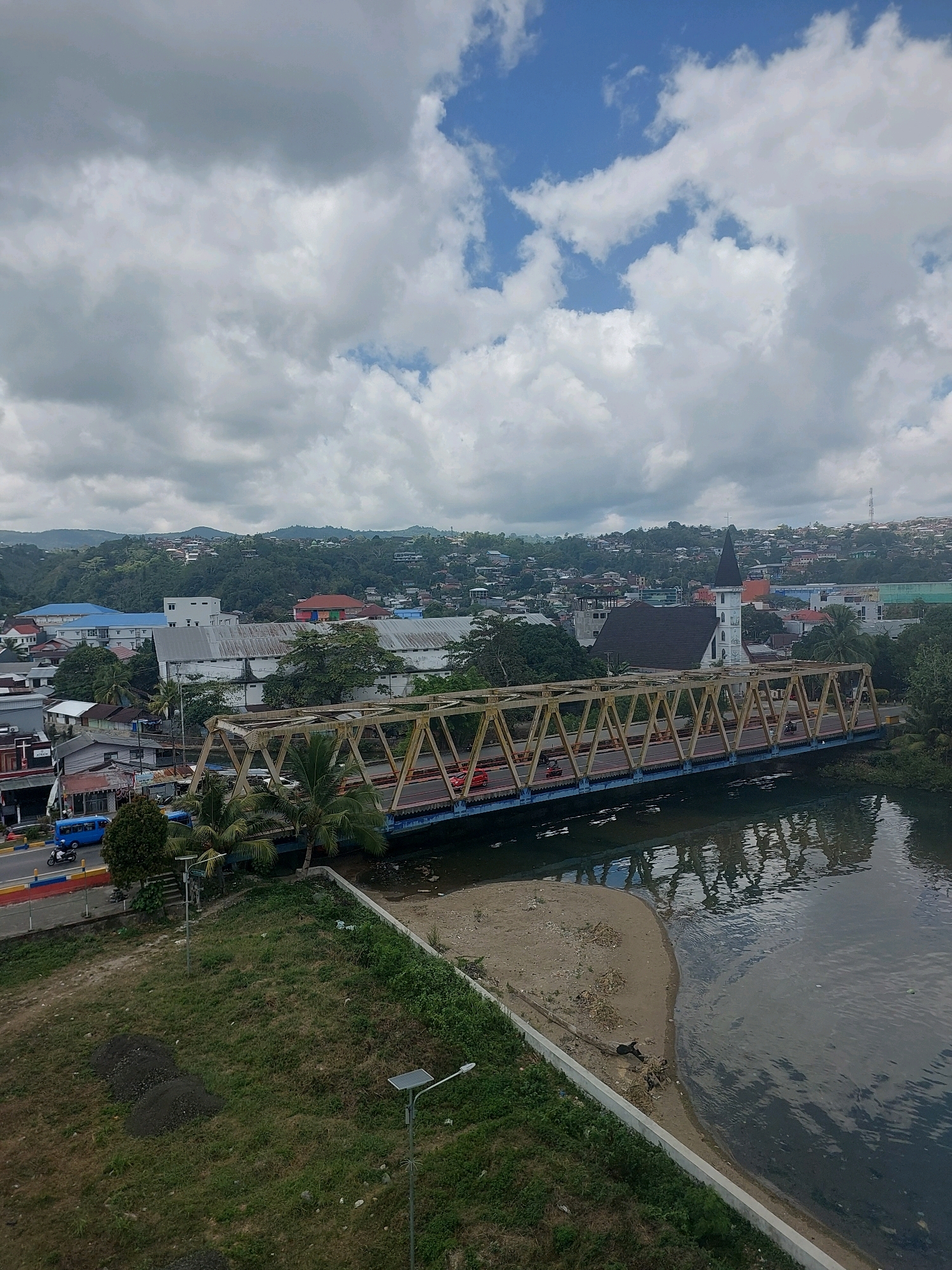 This aerial, overcast image depicts a view over the Batumerah River in Ambon, Indonesia. A large, yellow truss bridge spans the river, carrying vehicular traffic. The bridge's structure is visible as a series of interconnected triangular beams painted yellow, with a blue railing and roadway. Several vehicles, including cars and motorcycles, are present on the bridge.
The river itself is dark and reflective, with the bridge's structure casting a distorted reflection on its surface. The riverbanks are a mix of sandy areas, vegetation, and concrete retaining walls. On the left bank, a grassy area with a few small mounds of dark soil and solar-powered streetlights is visible.
Beyond the bridge and river, the cityscape of Ambon unfolds. Buildings of various sizes and colors are clustered together, extending up the rolling green hills that form the backdrop of the scene. Prominent structures include a white building with a large metal roof, and a white church with a black steeple. Residential housing is spread across the hillsides. The sky is filled with a dramatic display of clouds, with patches of blue sky visible. The overall impression is of a densely populated urban area nestled within a hilly, tropical landscape.