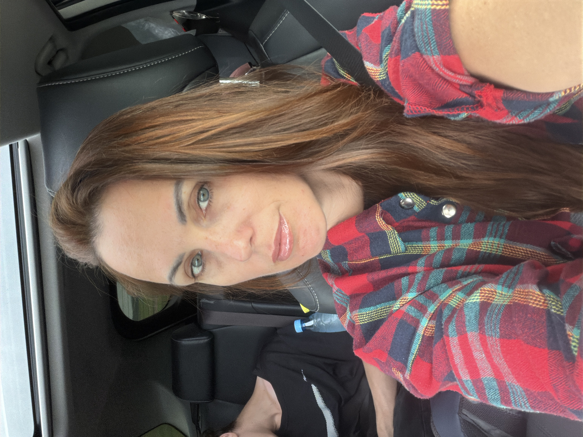 In an outdoor, daytime shot, a woman with long, reddish-brown hair and blue eyes is taking a selfie from the driver's seat of a car. She is wearing a red, blue, and yellow plaid shirt. Her lips are glossy and light pink. In the background, a person wearing a black t-shirt is visible in the passenger seat. The car's interior is dark leather, and a sunroof is open overhead. A water bottle is in the cup holder. The location is the city of Kirga, Russia.