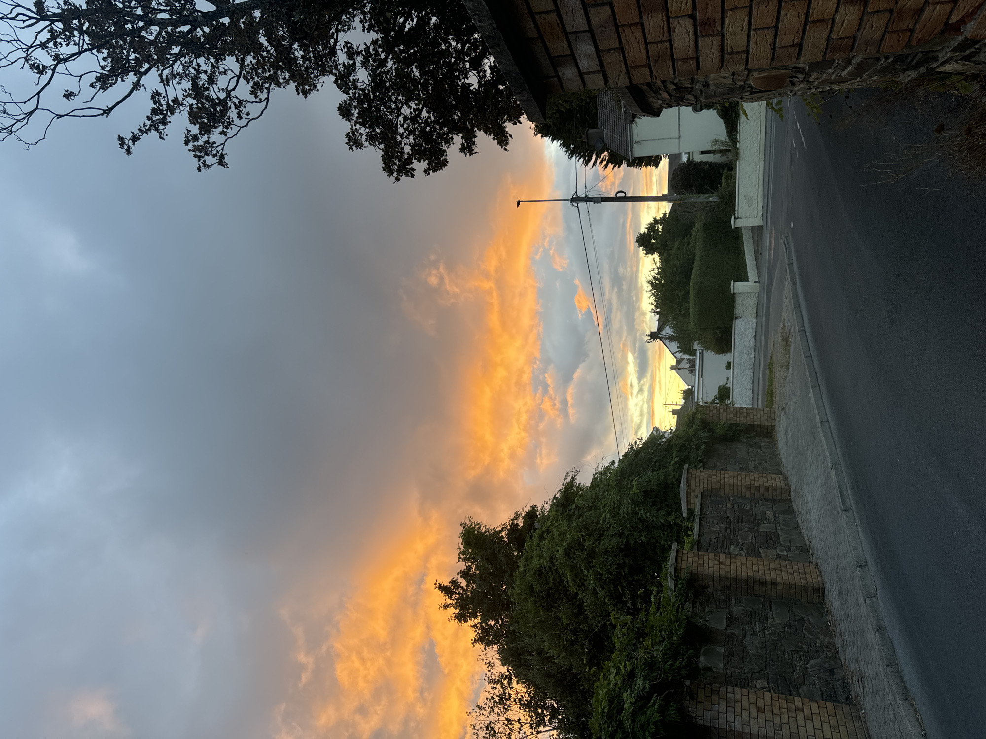The image depicts a street scene in Waterford, Ireland, at dusk. The sky is filled with dramatic clouds, glowing with hues of orange and yellow from the setting sun, contrasting with the darker, grey clouds above. To the left, a stone wall topped with brick pillars and lush green foliage lines the sidewalk. The road, paved with asphalt, curves gently to the right. In the background, houses with white walls and tiled roofs are visible amidst mature trees and manicured hedges. A tall street lamp stands on the right side of the road, silhouetted against the colorful sky. The overall impression is one of a quiet residential area experiencing a vibrant sunset.