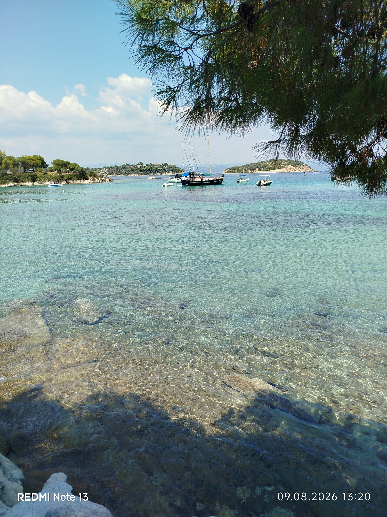 The image depicts a coastal scene in Sithonia Municipal Unit, Greece, likely a public beach or bay. In the foreground, a rocky shoreline is visible with clear, turquoise water lapping at the stones, allowing the seabed to be seen. Above the water, branches of a pine tree hang into the frame, casting shadows on the water.
In the middle ground and background, the expansive turquoise sea stretches out, dotted with several boats. One larger boat, appearing to be a sailing vessel with a dark hull and white sails, is anchored on the left. Several smaller motorboats are also present in the water.
The shoreline on the far side of the bay is lined with lush green trees and vegetation, with some buildings visible amongst the foliage, suggesting a populated coastal area or resort. The sky above is a clear blue with scattered white clouds. The lighting indicates it is daytime, likely late morning or early afternoon given the date and time stamp on the image.
The overall scene conveys a serene and picturesque coastal landscape, characteristic of the Sithonia region.