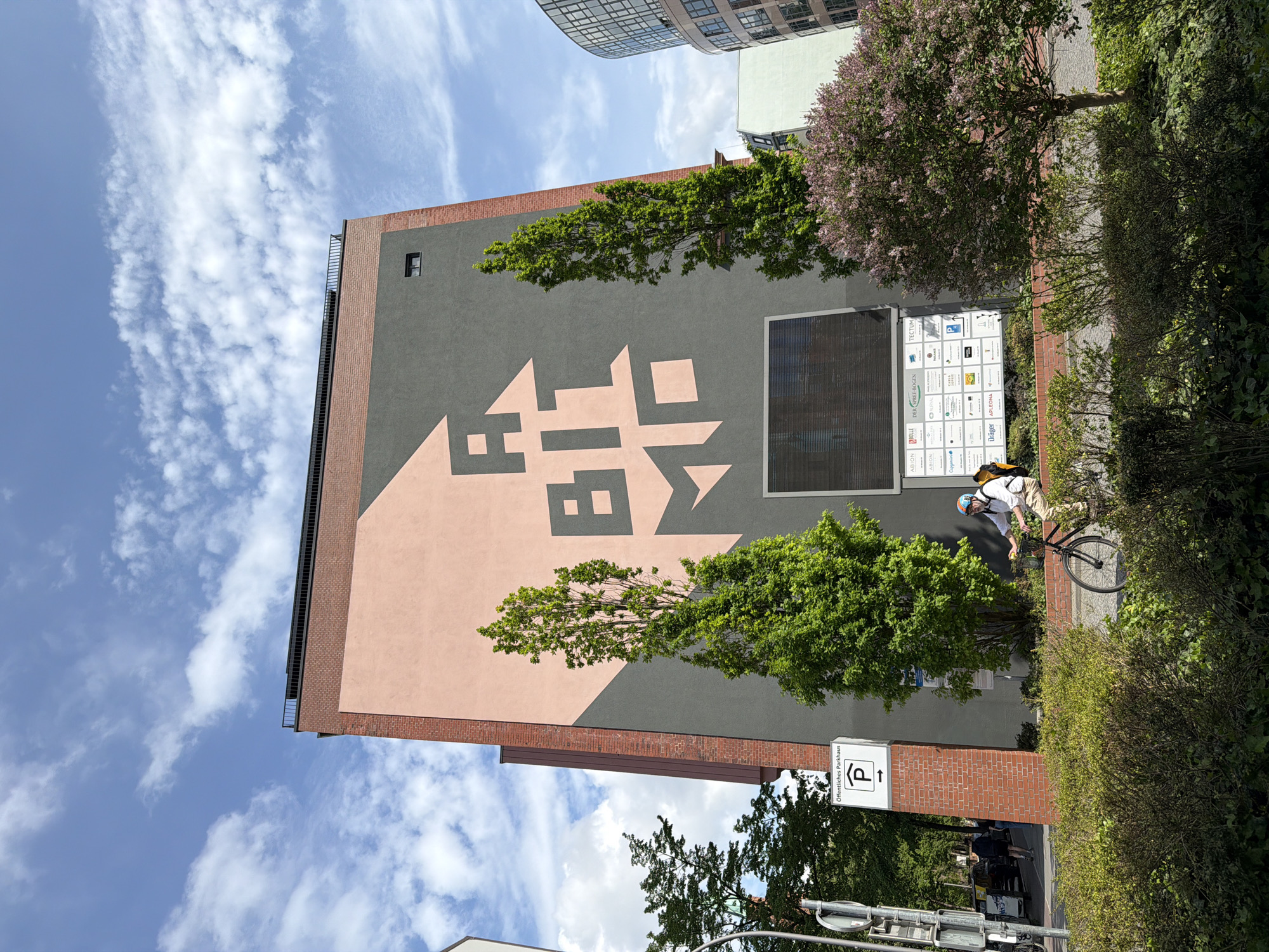The image shows the side of a building in Berlin, Germany, decorated with a large geometric design in shades of gray and peach. The upper portion of the building is gray, while the lower portion features the peach-colored geometric pattern. Two slim trees are positioned in front of the building's facade, partially obscuring the artwork. To the right of the trees, a man wearing a white shirt, khaki pants, and a helmet is seen riding a bicycle on the sidewalk. A sign indicating a public parking garage ("Öffentliches Parkhaus") is visible next to the sidewalk. A display board with company logos, including "TECTUM", "APLEONA" and "Capgemini," is also affixed to the building's facade. The sky above is blue with scattered clouds.