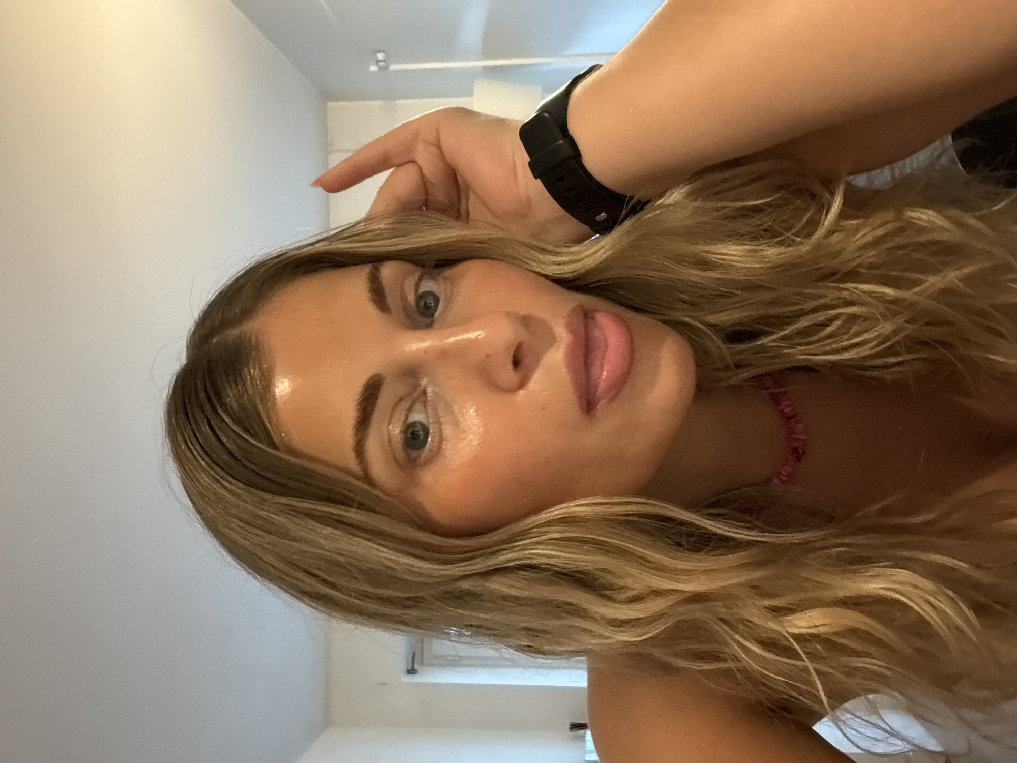 A close-up, rotated view depicts a woman with blonde, wavy hair and fair skin. She has a black watch on her wrist, a pink beaded necklace, and appears to be in an interior setting with white walls and a window in the background. There is no evidence of the image being taken in Paris, France.